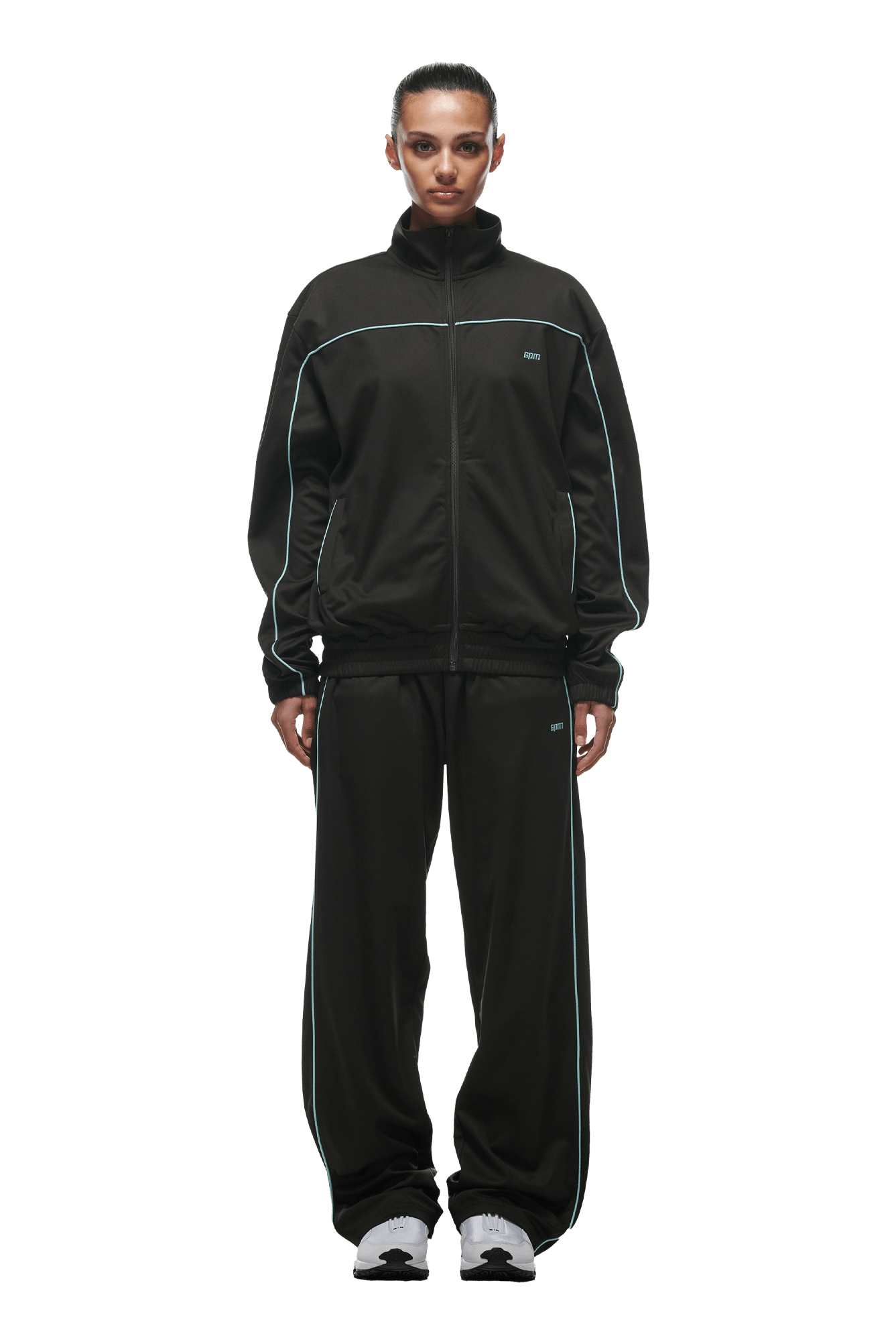 A person stands facing forward wearing the 6PM 6PM+ TRAINER JACKET ANTHRACITE with white piping and zipped pockets, paired with white sneakers and slicked-back hair, against a solid green background.