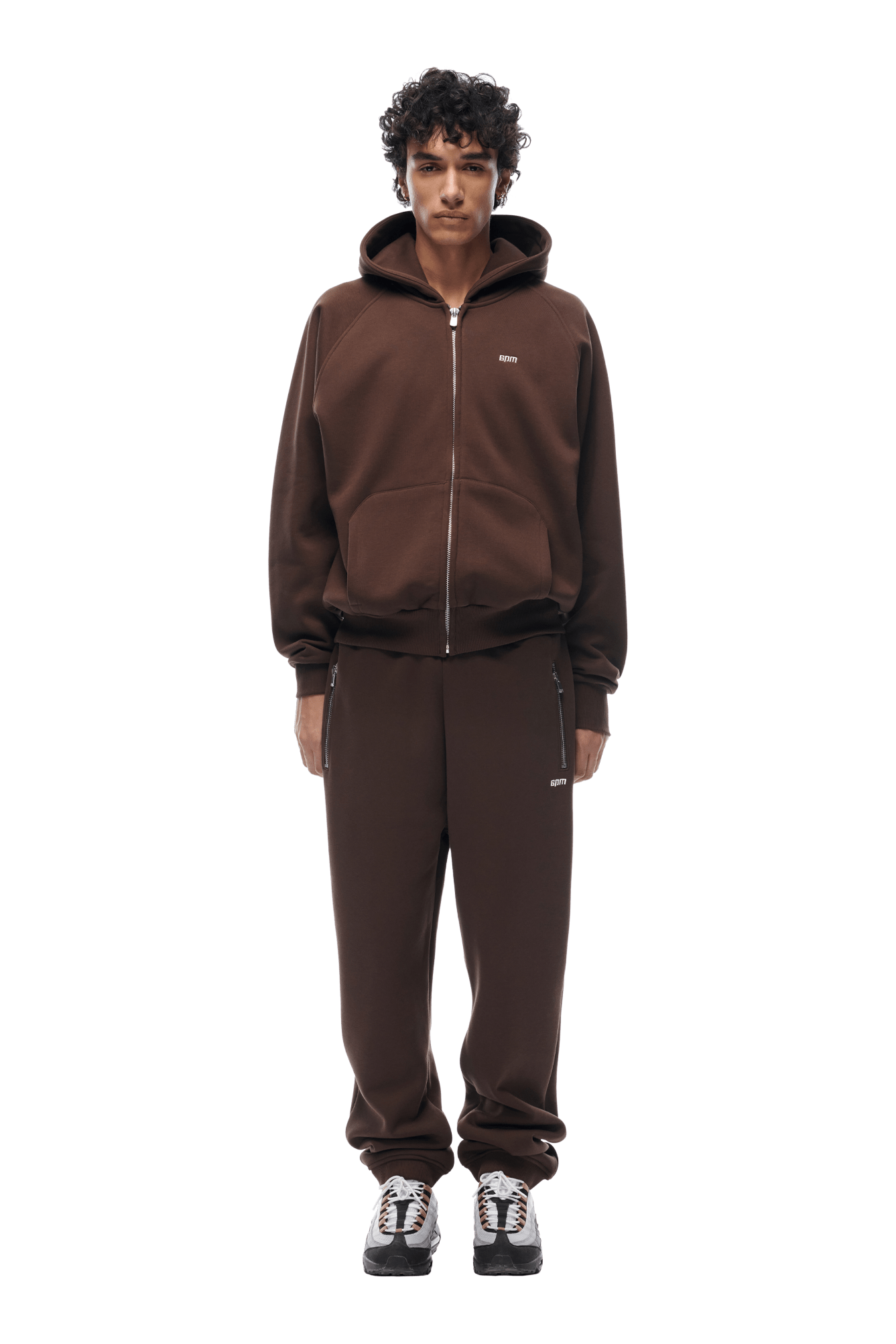 A person with curly hair wears the 6PM ZIP HOODIE CHOCOLATE BROWN and matching sweatpants, paired with white and black sneakers. The clean, everyday look stands out against a transparent background.
