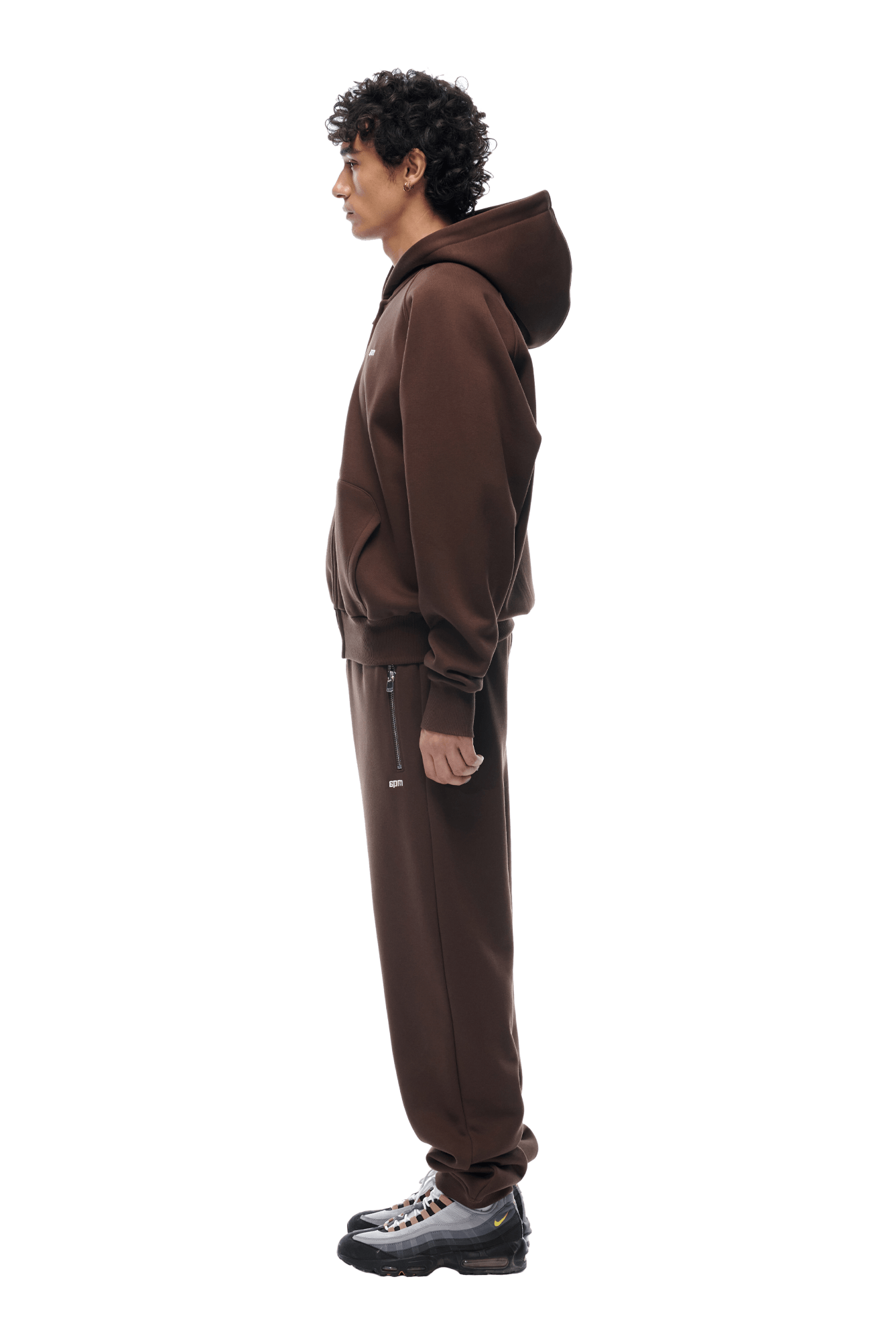 A person with curly hair stands in profile, wearing the 6PM ZIP HOODIE CHOCOLATE BROWN and matching sweatpants with black sneakers against a plain background—its clean finish makes it perfect for everyday wear.