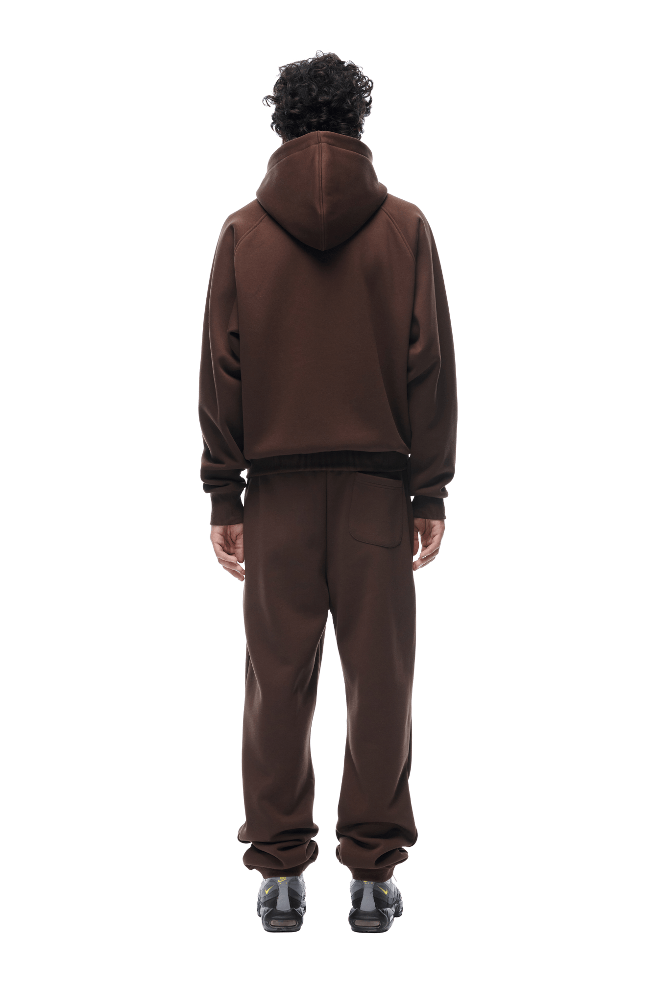 A person with curly hair, seen from behind, wears the 6PM ZIP HOODIE CHOCOLATE BROWN and matching sweatpants with black sneakers on a transparent background—a comfortable, stylish everyday look.