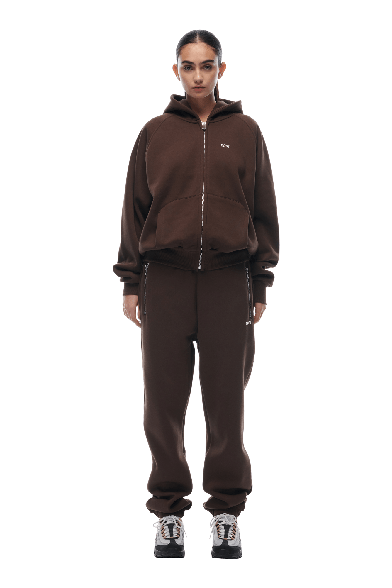 A person stands facing forward in the 6PM ZIP HOODIE CHOCOLATE BROWN set, paired with matching joggers and black-and-white sneakers. With slicked-back dark hair and hands by their sides, they present a clean look against a transparent background.
