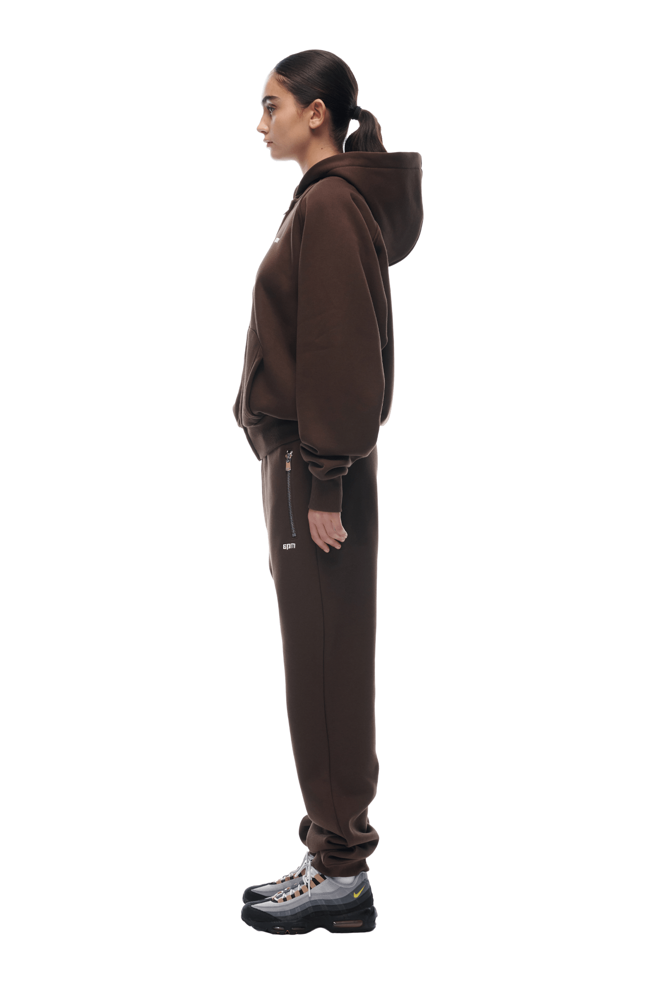 A woman stands in profile with hair in a ponytail, wearing the 6PM ZIP HOODIE CHOCOLATE BROWN and matching pants, paired with silver and black sneakers. Outfit shown on a transparent background, ideal for everyday wear.