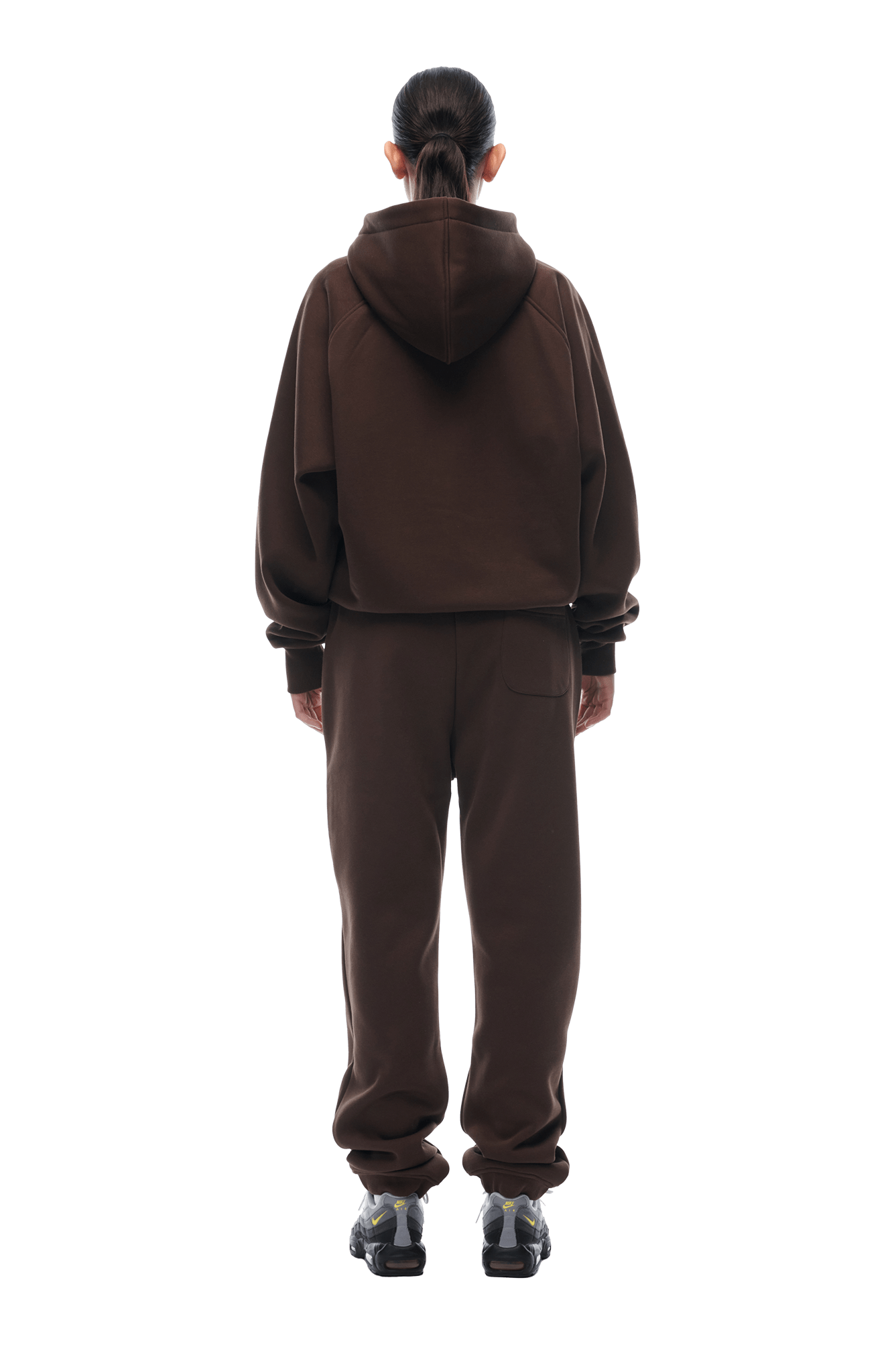 A person with long dark hair in a ponytail faces away, wearing the 6PM ZIP HOODIE CHOCOLATE BROWN with matching sweatpants and black sneakers, set against a plain background.