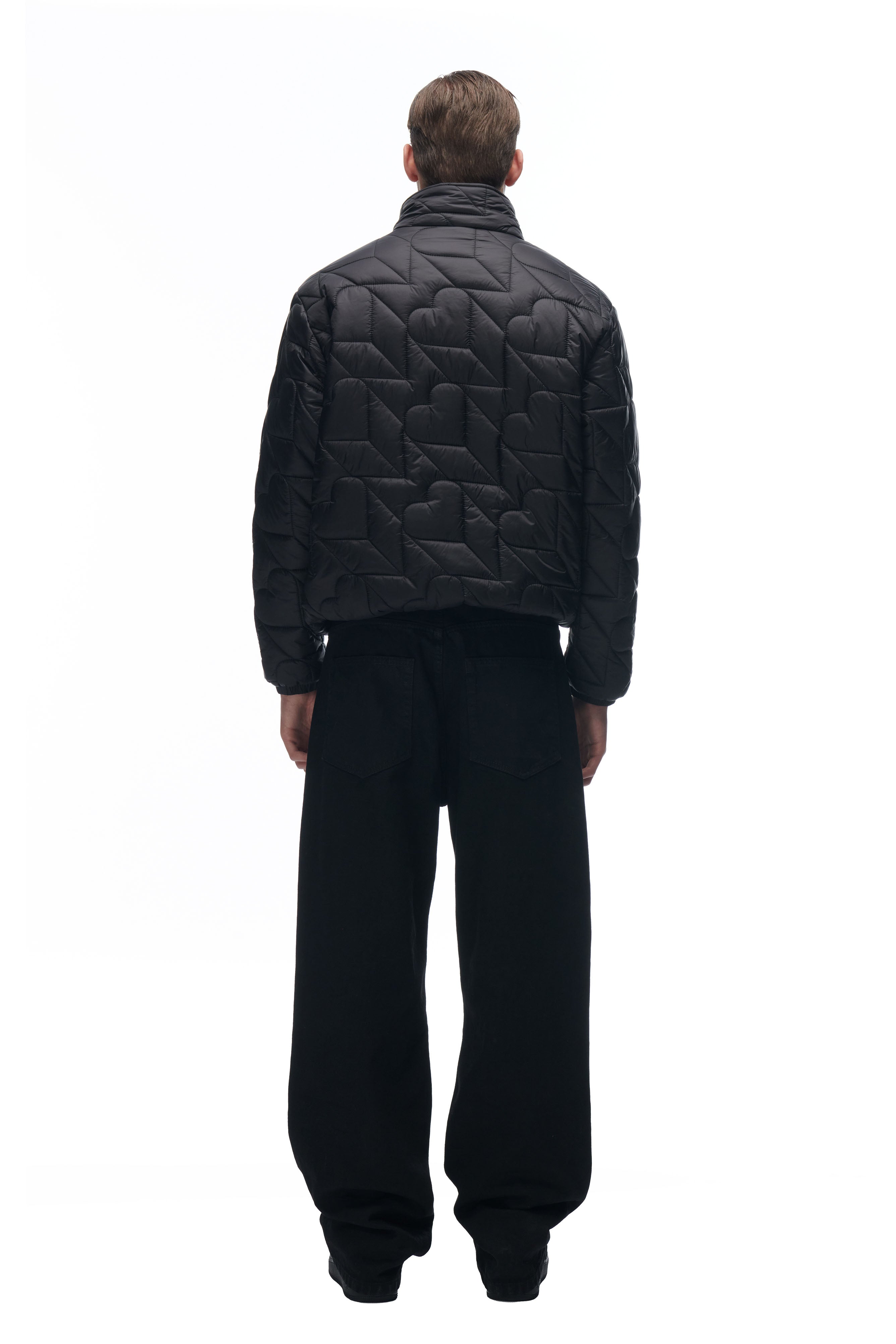 A person faces away, wearing the 6PM HEART SPRING JACKET—a black quilted jacket with a heart pattern—paired with loose black pants, set against a plain white background.