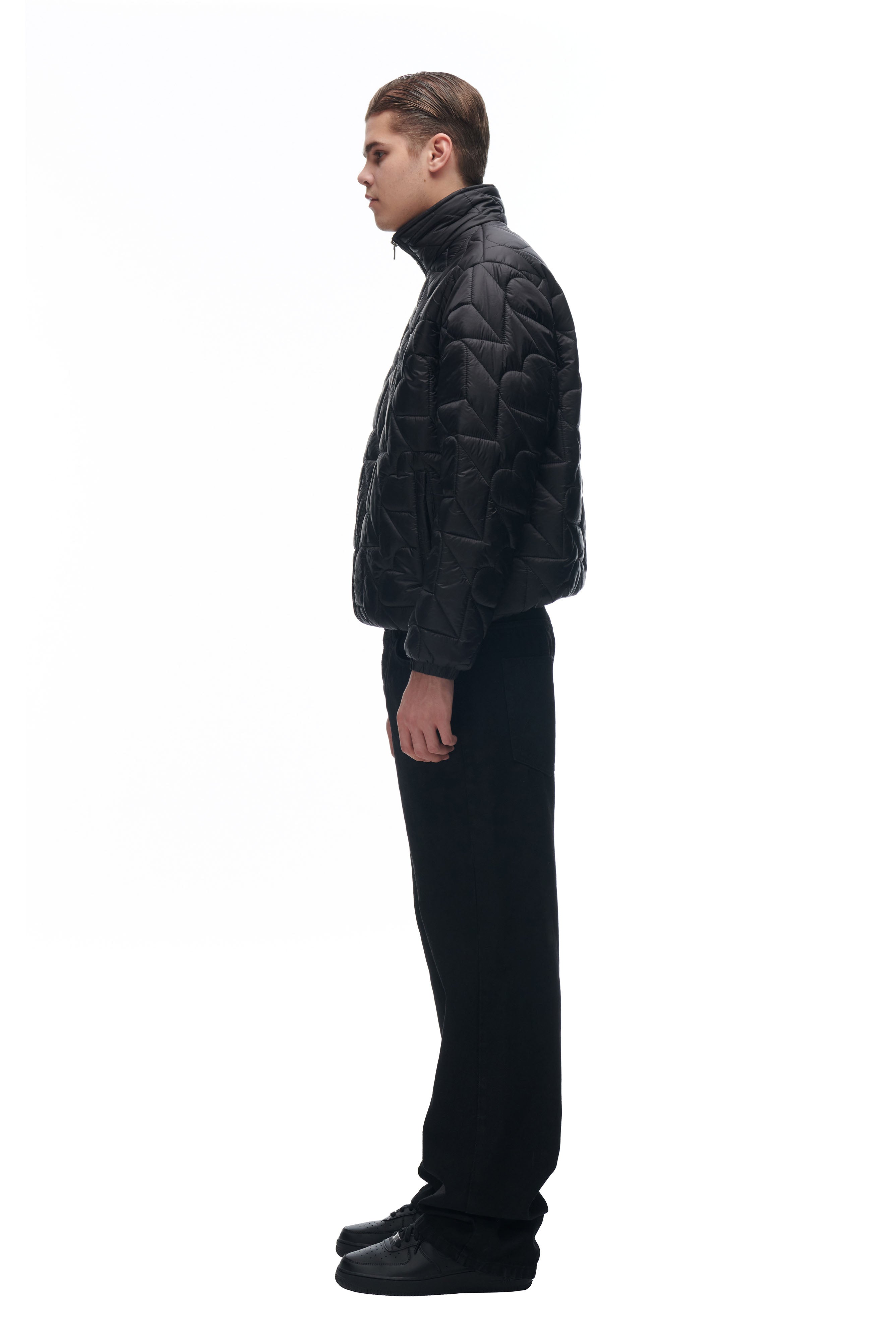 A person in profile faces a white background, wearing the 6PM HEART SPRING JACKET with a high collar, paired with black pants and shoes. Their hair is short and slicked back.
