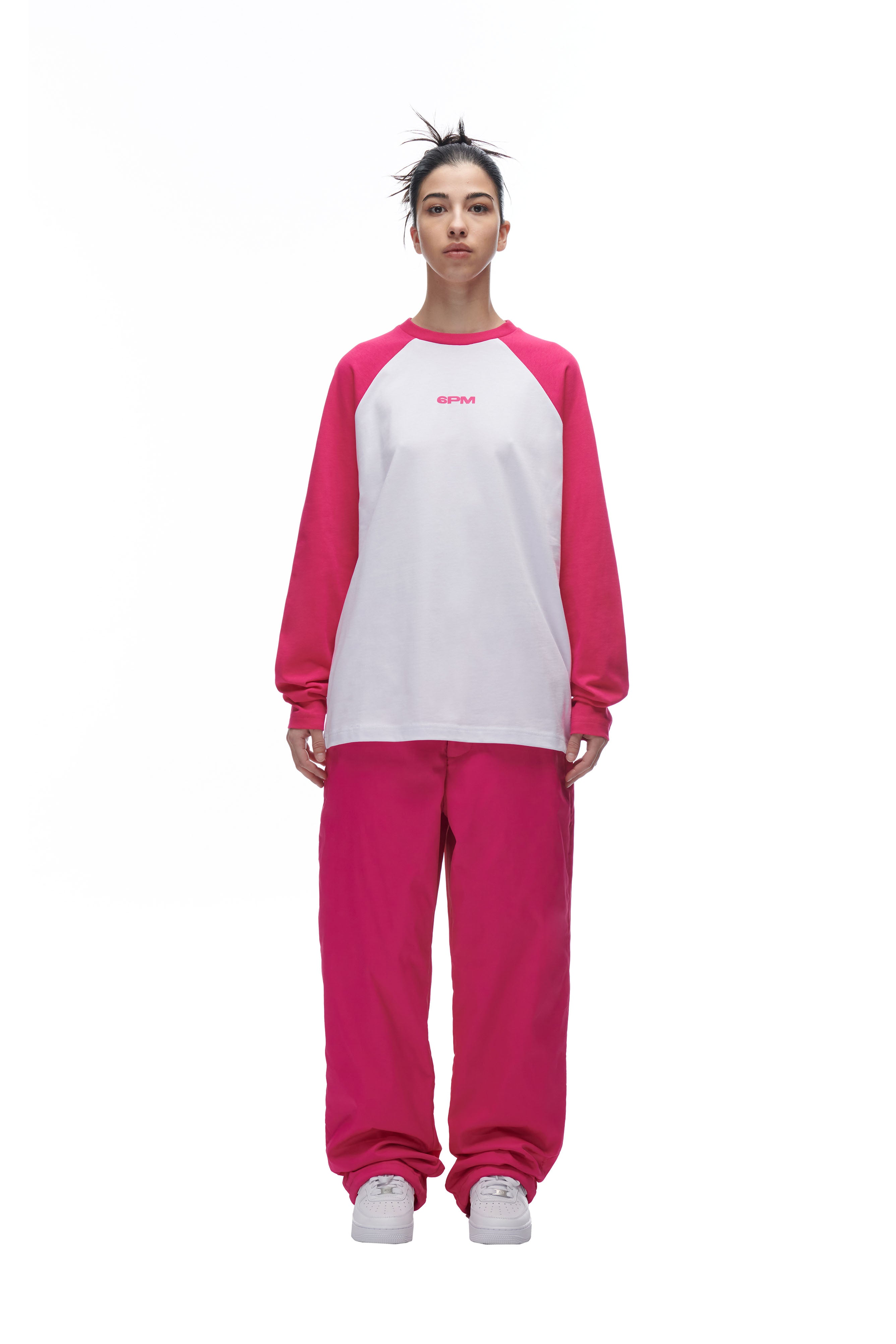 A person faces forward in a 6PM LH LONGSLEEVE WHITE/PINK shirt with bright pink sleeves, matching pink pants, light gray sneakers, and messy bun hair, all set against a plain white background.