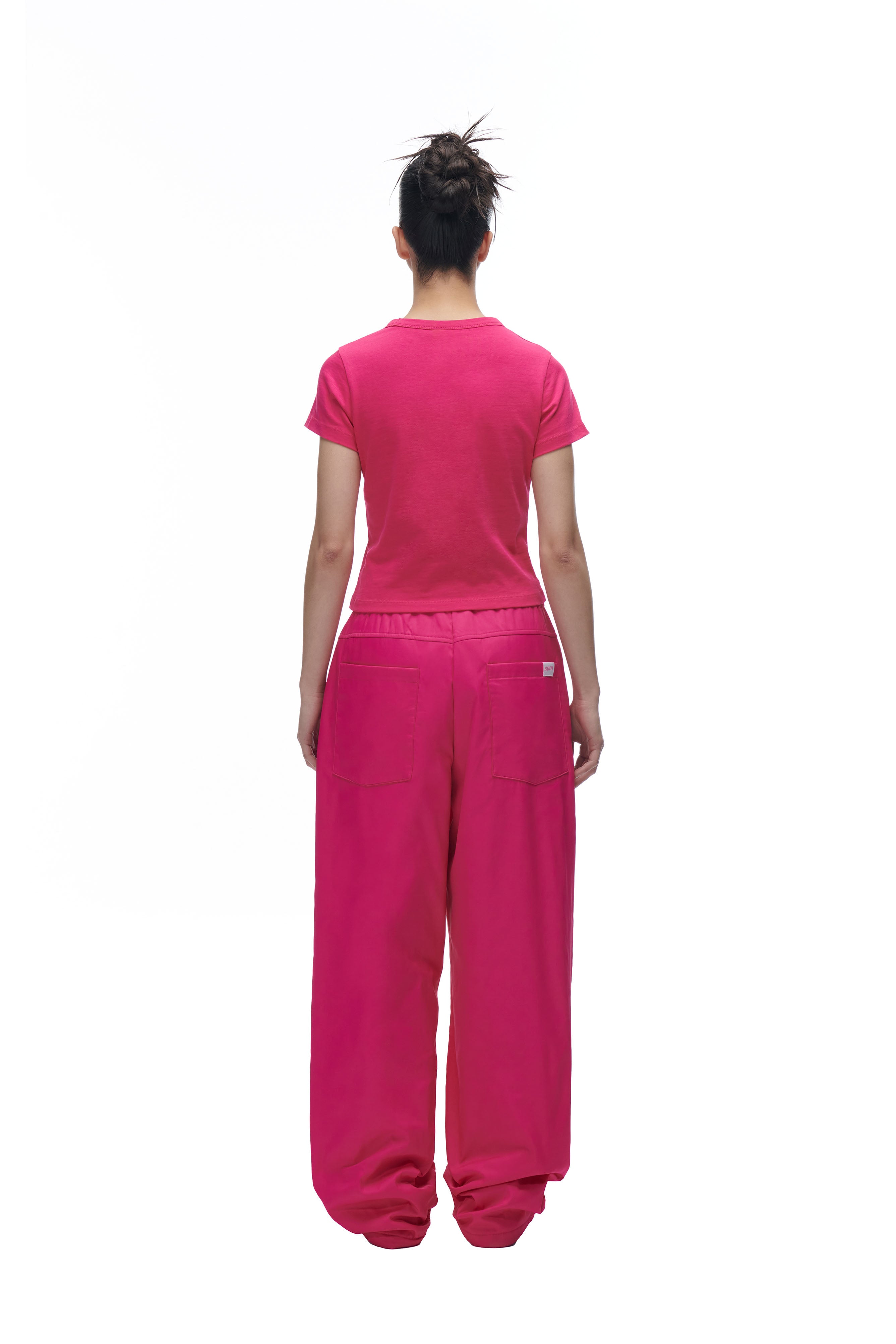A person with their hair in a messy bun is standing facing away, wearing the 6PM LOGO BABYTEE PINK and loose-fitting pink pants against a white background.
