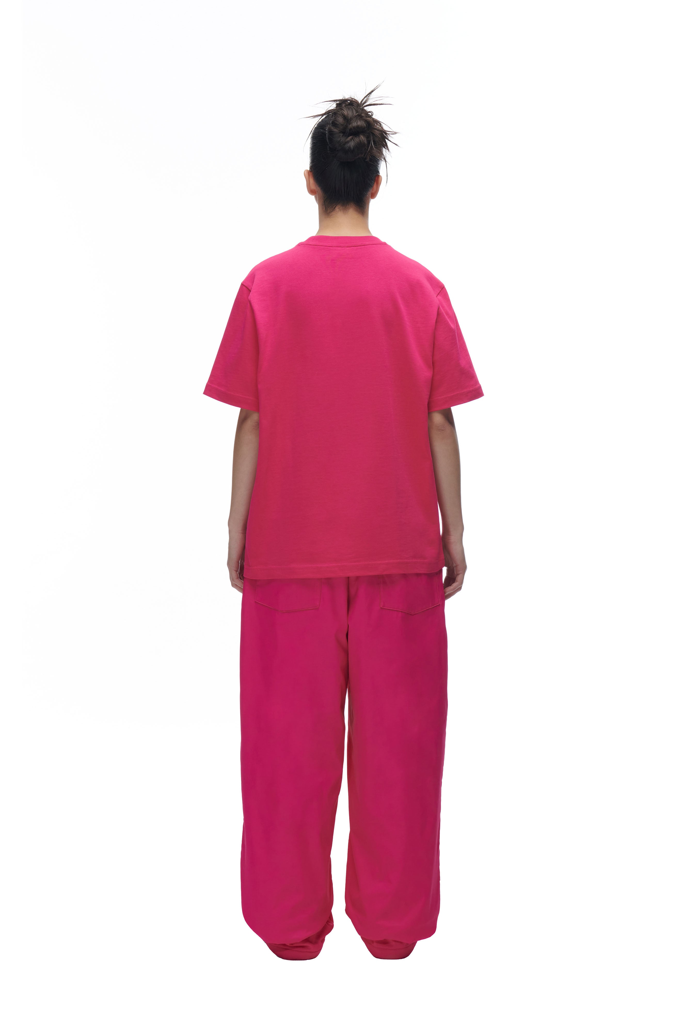 Model facing away from the camera wears the 6PM DATENIGHT T-SHIRT PINK with matching wide pink pants. Hair is styled in a messy bun against a plain white background.