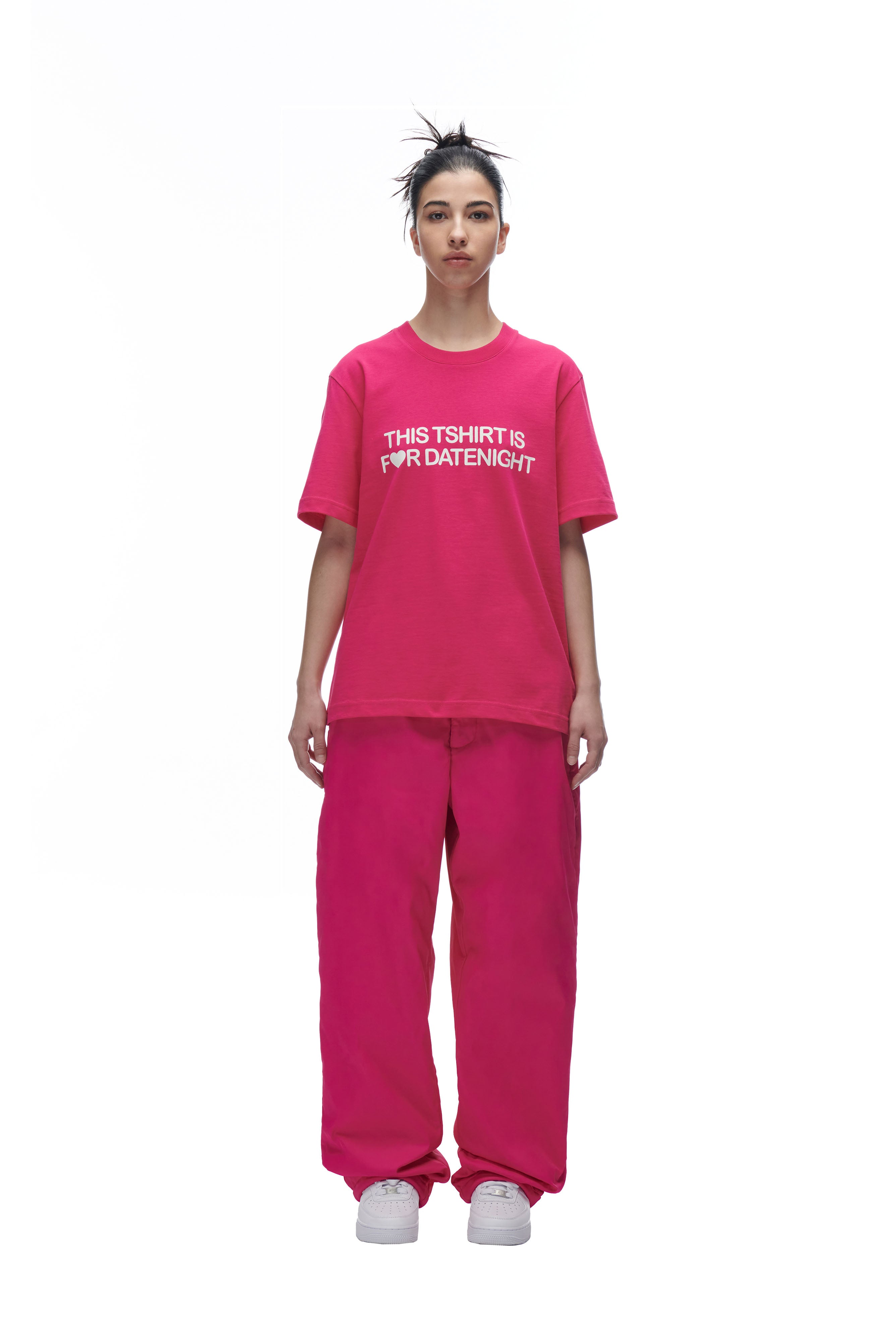 A person stands facing forward in a 6PM DATENIGHT T-SHIRT PINK, loose pink pants, and white sneakers, hair in a messy bun, against a white background.