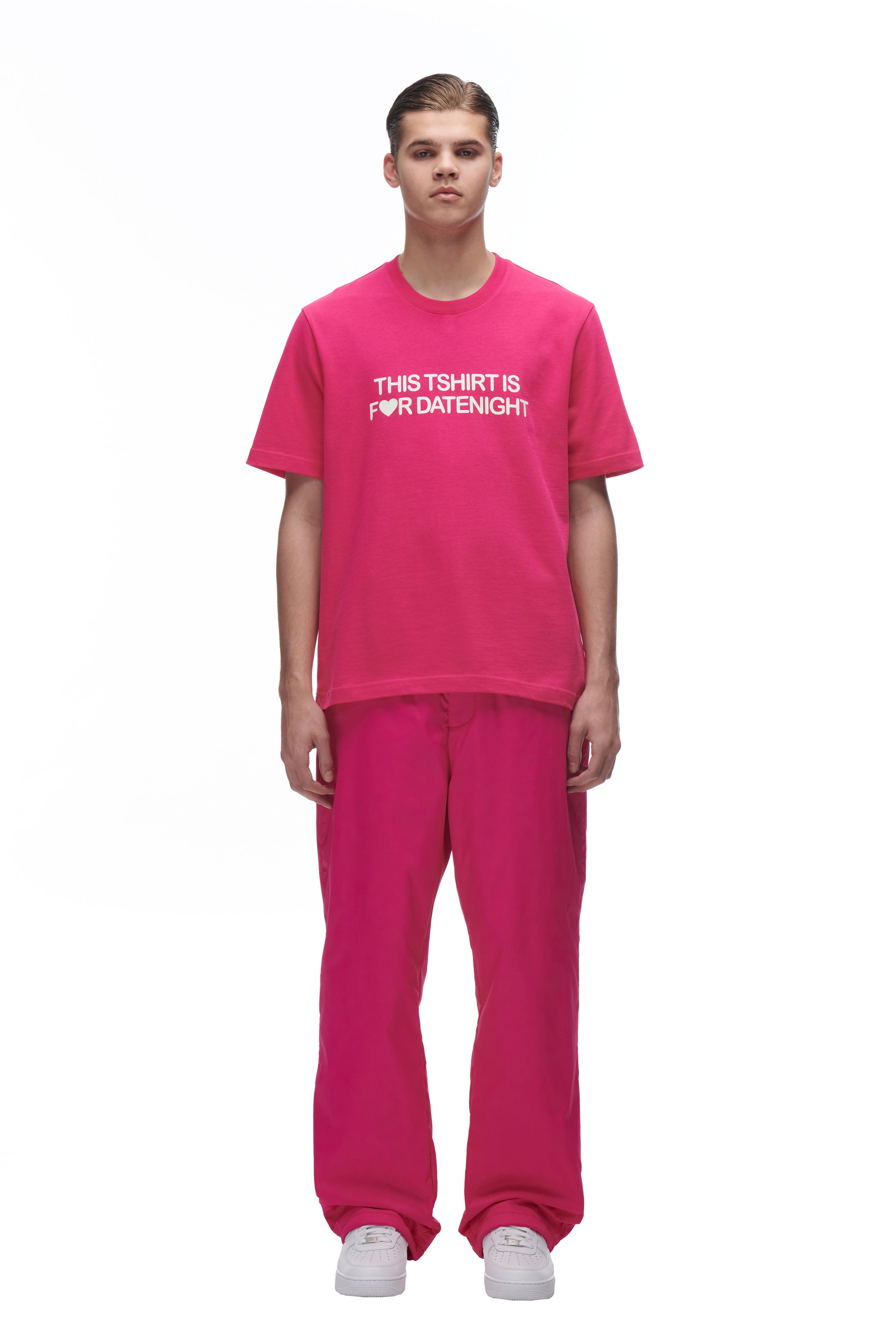 A person stands facing forward, wearing the 6PM DATENIGHT T-SHIRT PINK with "THIS TSHIRT IS FOR DATENIGHT" in white text, pink pants, and white sneakers against a plain white background.