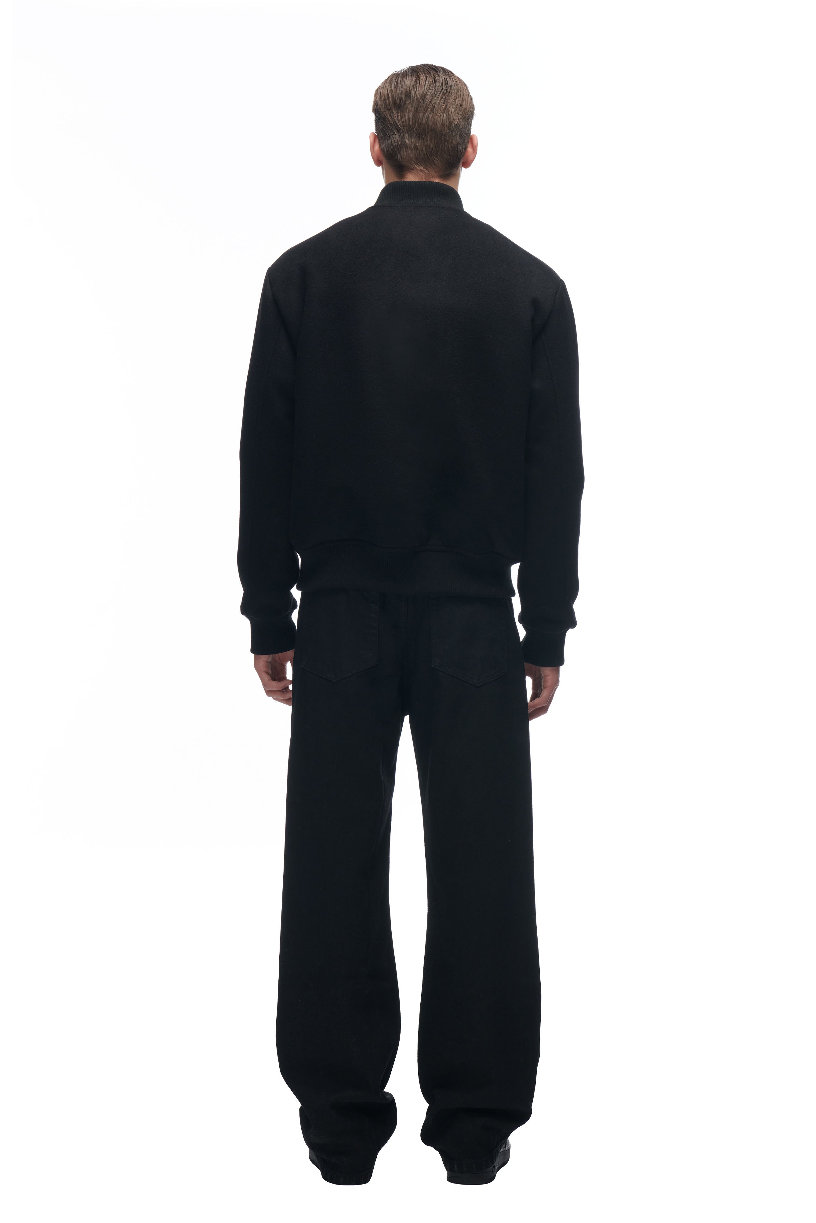 A person stands facing away from the camera, wearing the 6PM LH VARSITY JACKET and loose black pants against a plain white background.