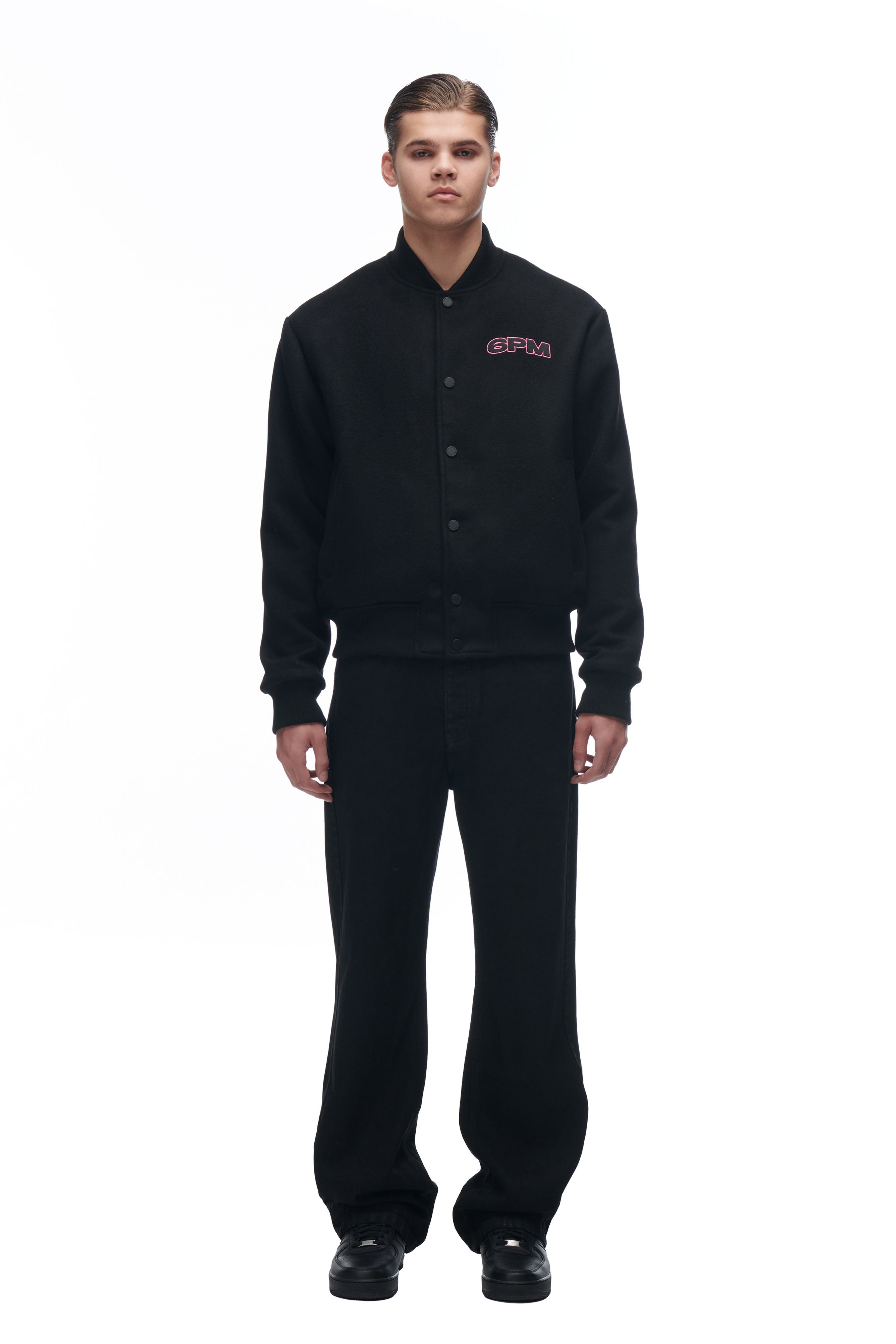 A person stands facing forward against a white background, wearing the 6PM LH VARSITY JACKET with a pink logo on the left chest, along with black pants and black shoes.