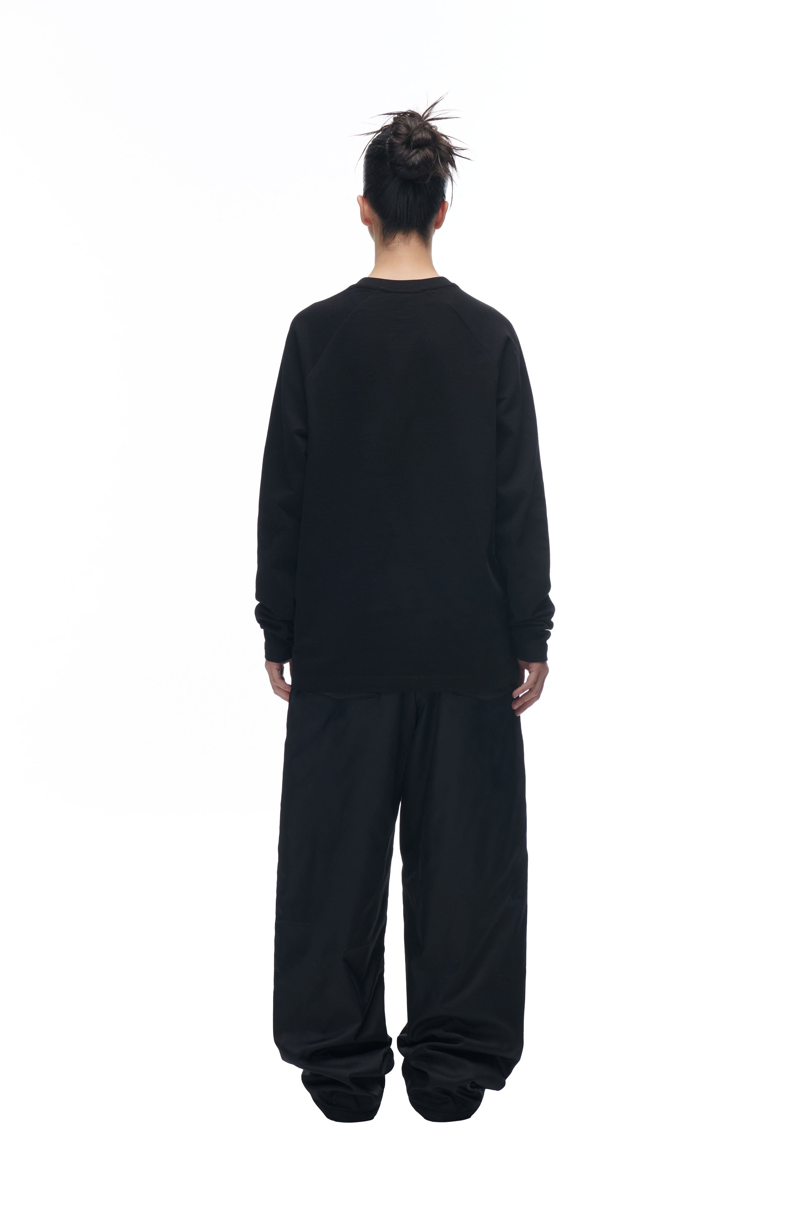Facing away against a white background, a person with dark hair in a messy bun wears the oversized 6PM LH LONGSLEEVE BLACK/BLACK shirt and wide-leg black pants.