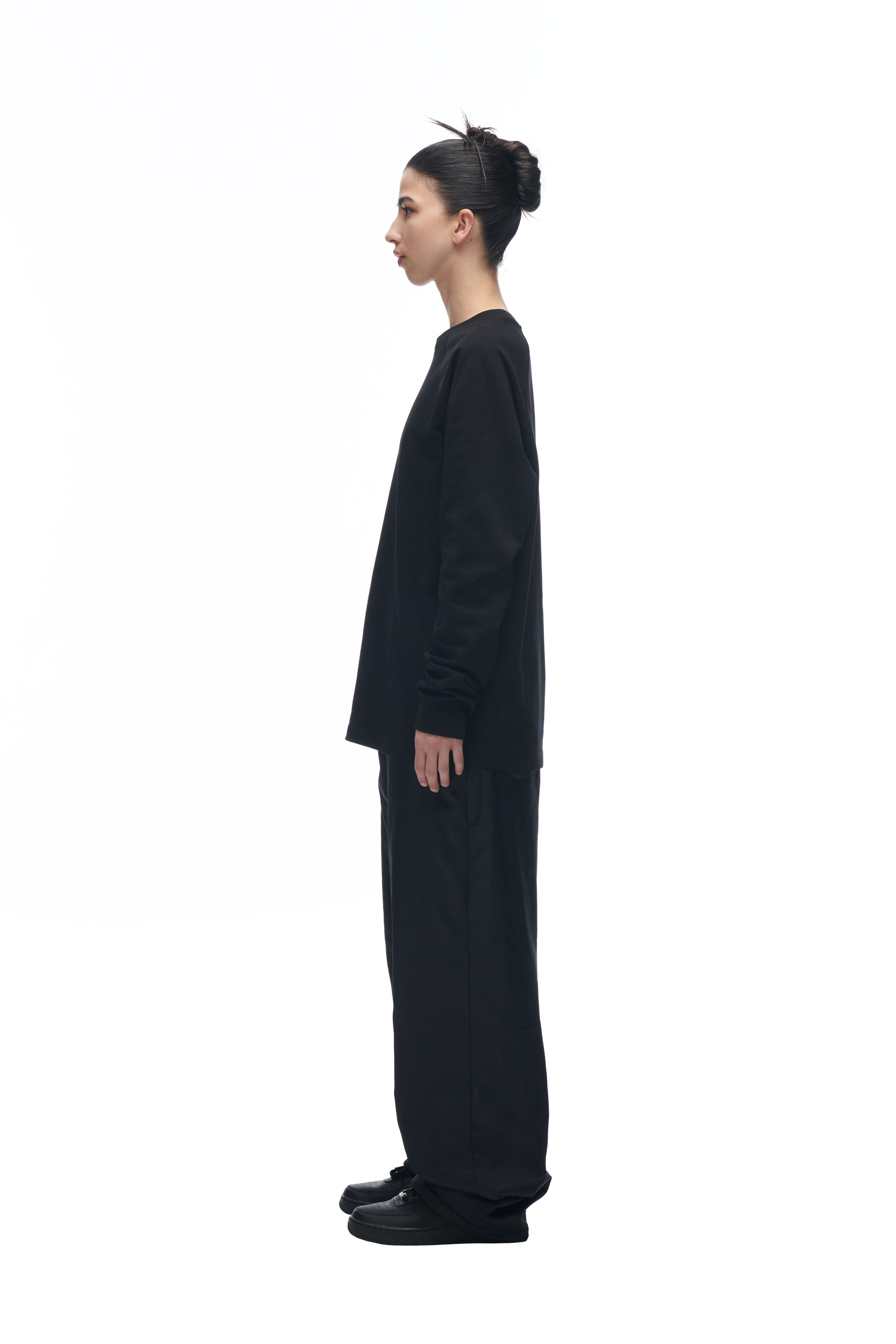 A person with dark hair in a bun wears the 6PM LH LONGSLEEVE BLACK/BLACK top and wide black pants, standing in profile against a white background.
