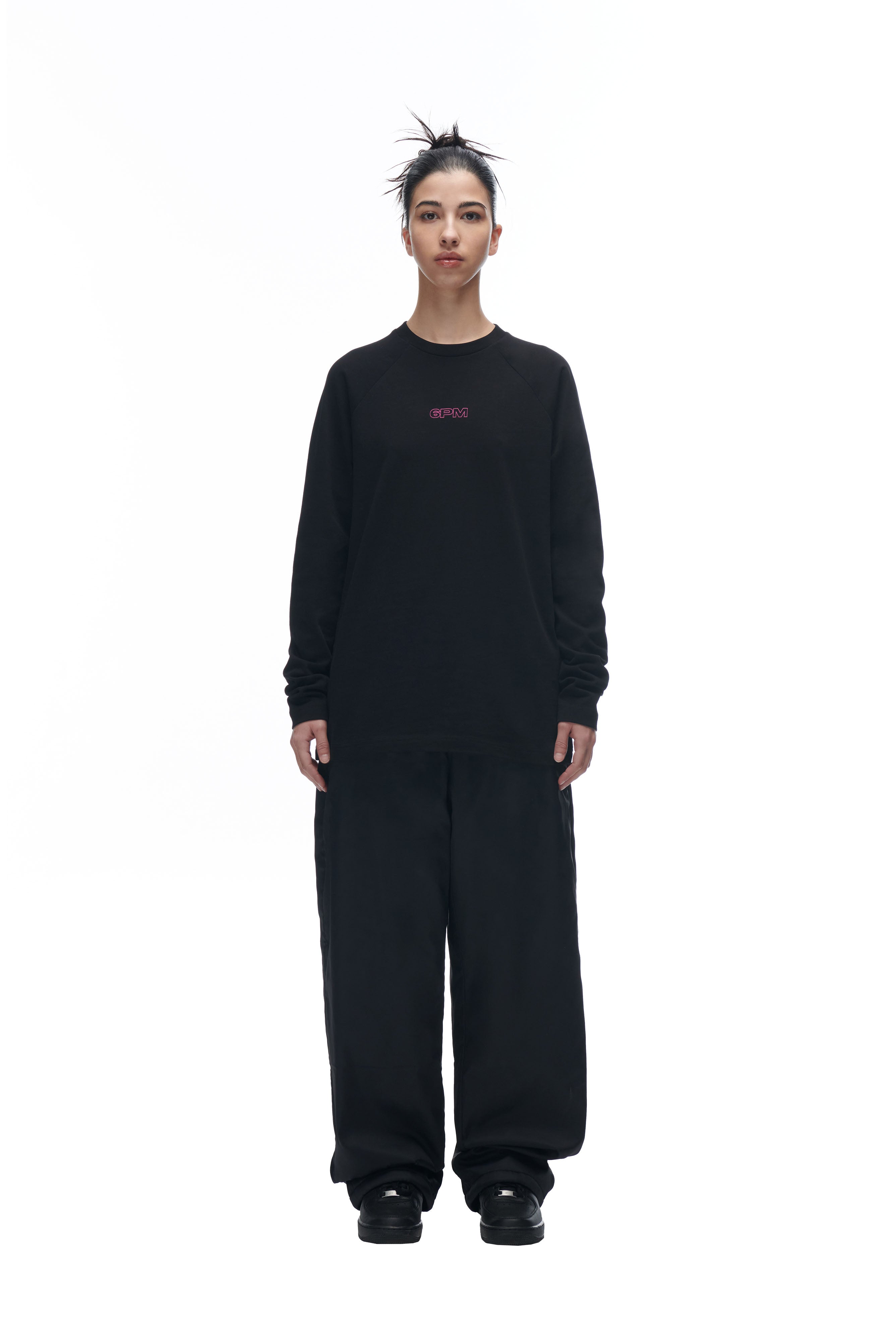 A person with a spiky updo and neutral expression stands against a white background, wearing the 6PM LH LONGSLEEVE BLACK/BLACK and loose black pants.