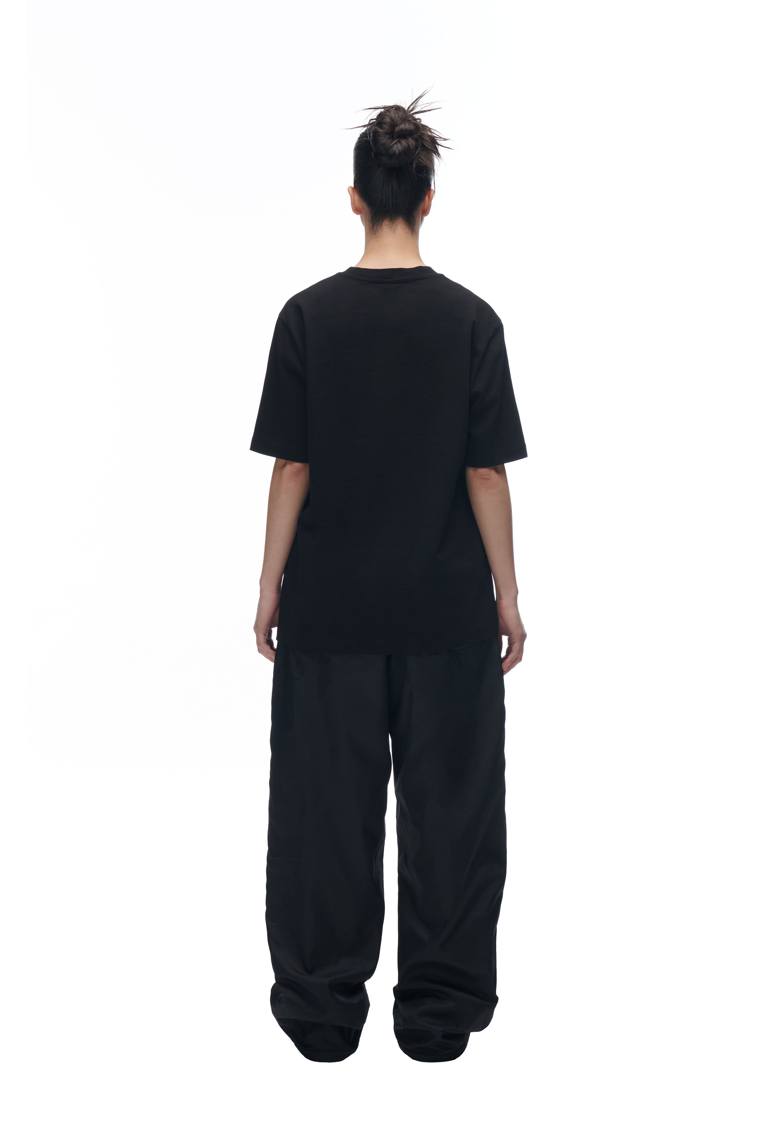 A person with their hair in a messy bun faces away, wearing the 6PM DATENIGHT T-SHIRT BLACK and baggy black pants against a plain white background.