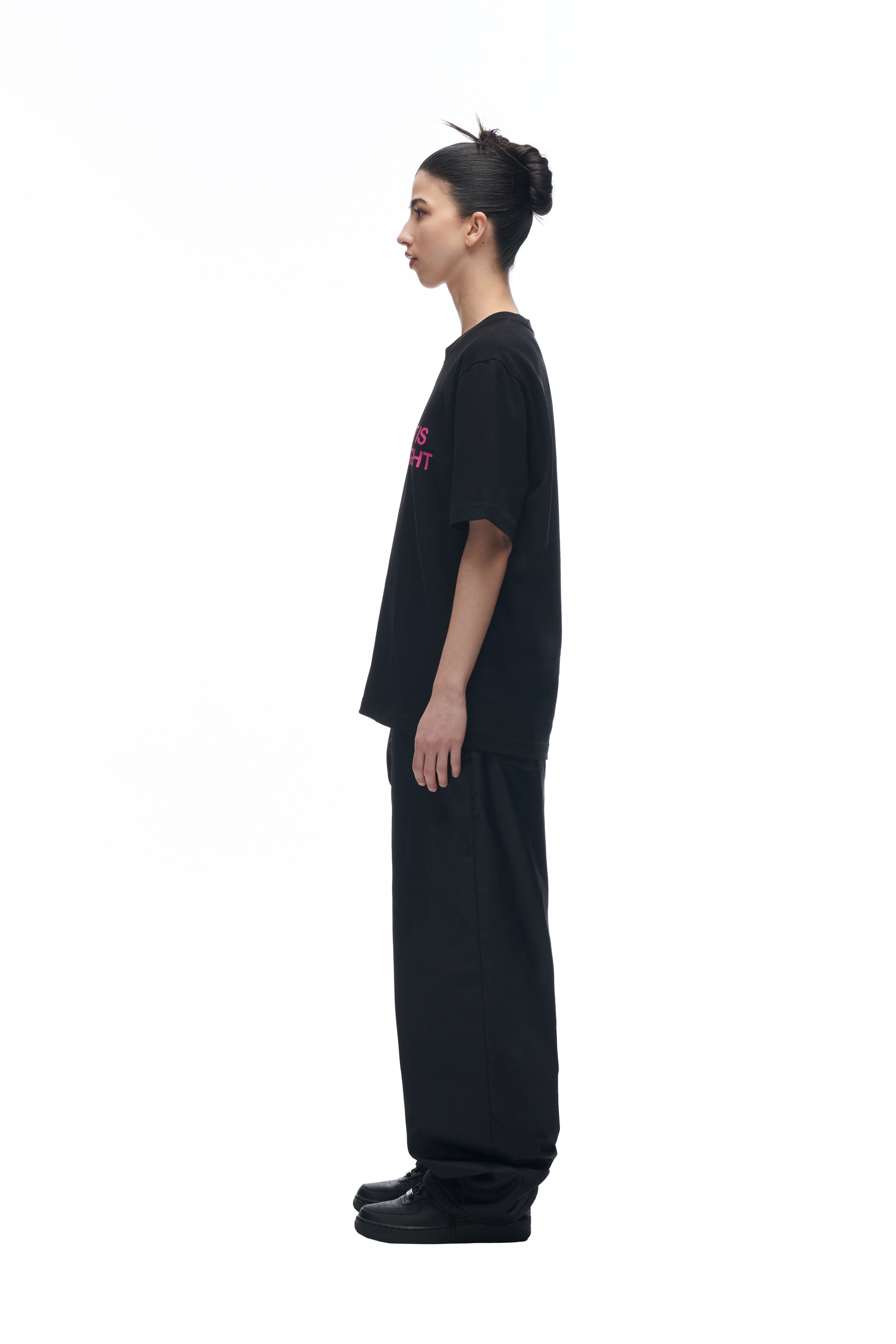 A person with dark hair in a bun stands in profile against a white background, wearing the 6PM DATENIGHT T-SHIRT BLACK, wide black pants, and black shoes.