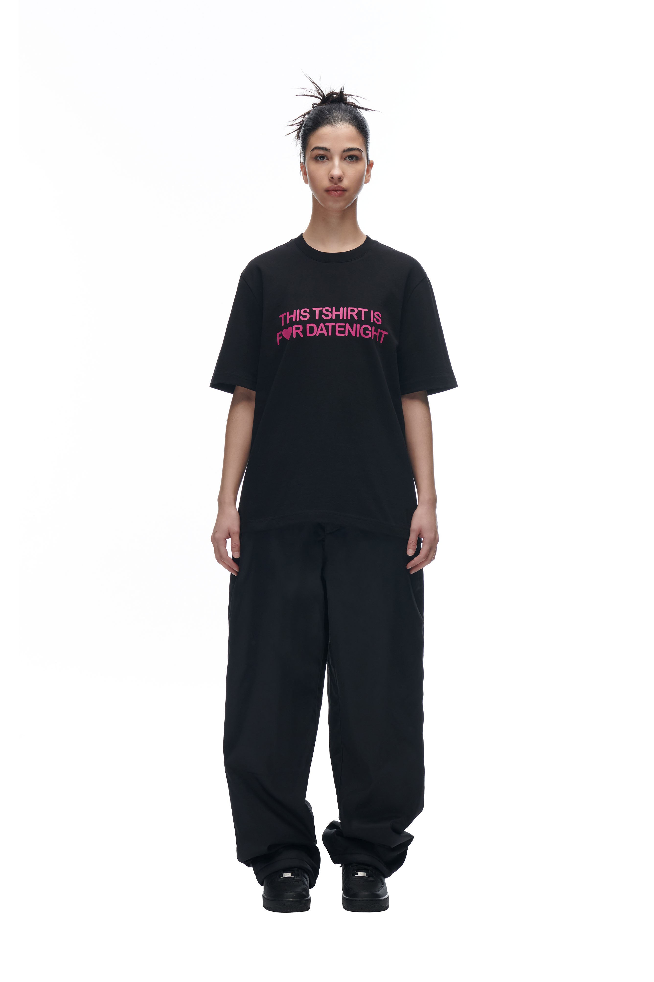 A person faces forward against a plain white background, wearing the 6PM DATENIGHT T-SHIRT BLACK—an oversized black tee with pink "THIS TSHIRT IS FOR DATENIGHT" text—styled with loose black pants and black shoes.
