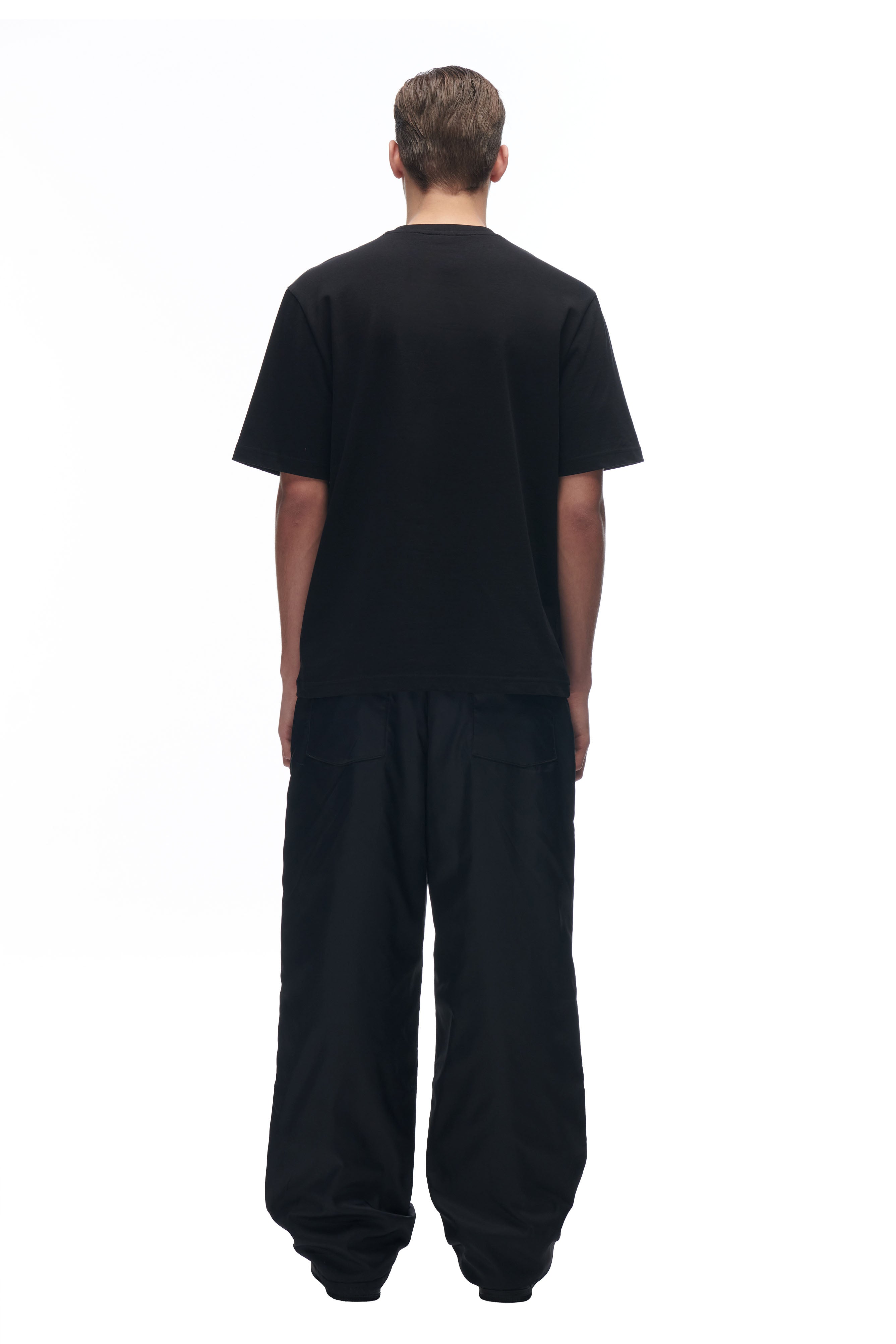 A person stands facing away from the camera in a 6PM DATENIGHT T-SHIRT BLACK and loose-fitting black pants, set against a white background.