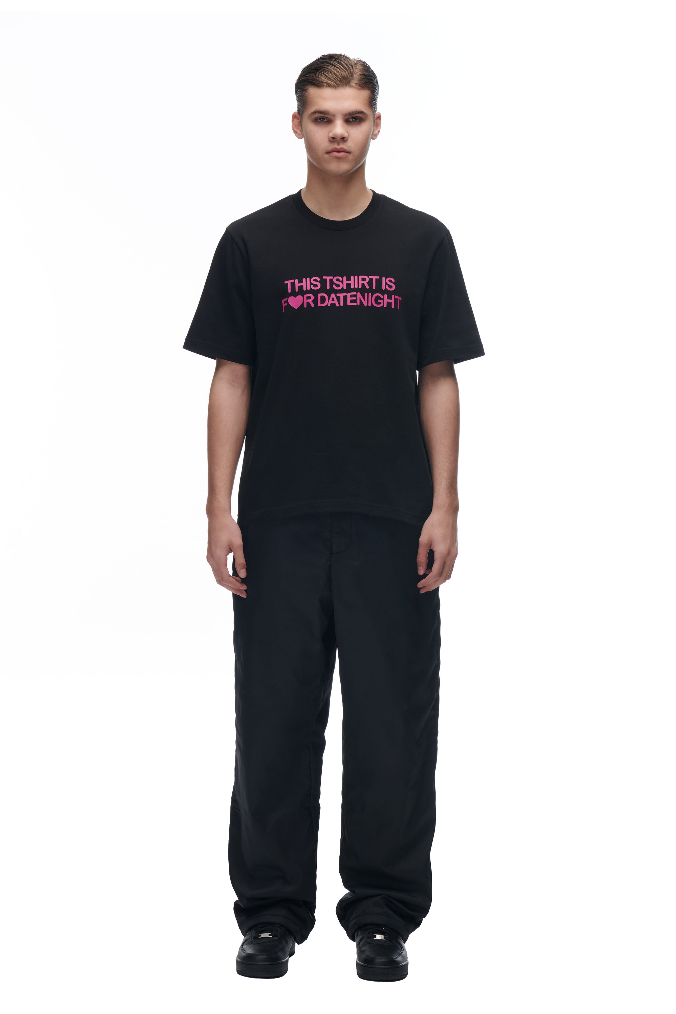 A young man faces forward wearing the 6PM DATENIGHT T-SHIRT BLACK with pink text, paired with loose black pants and black shoes, against a plain white background.
