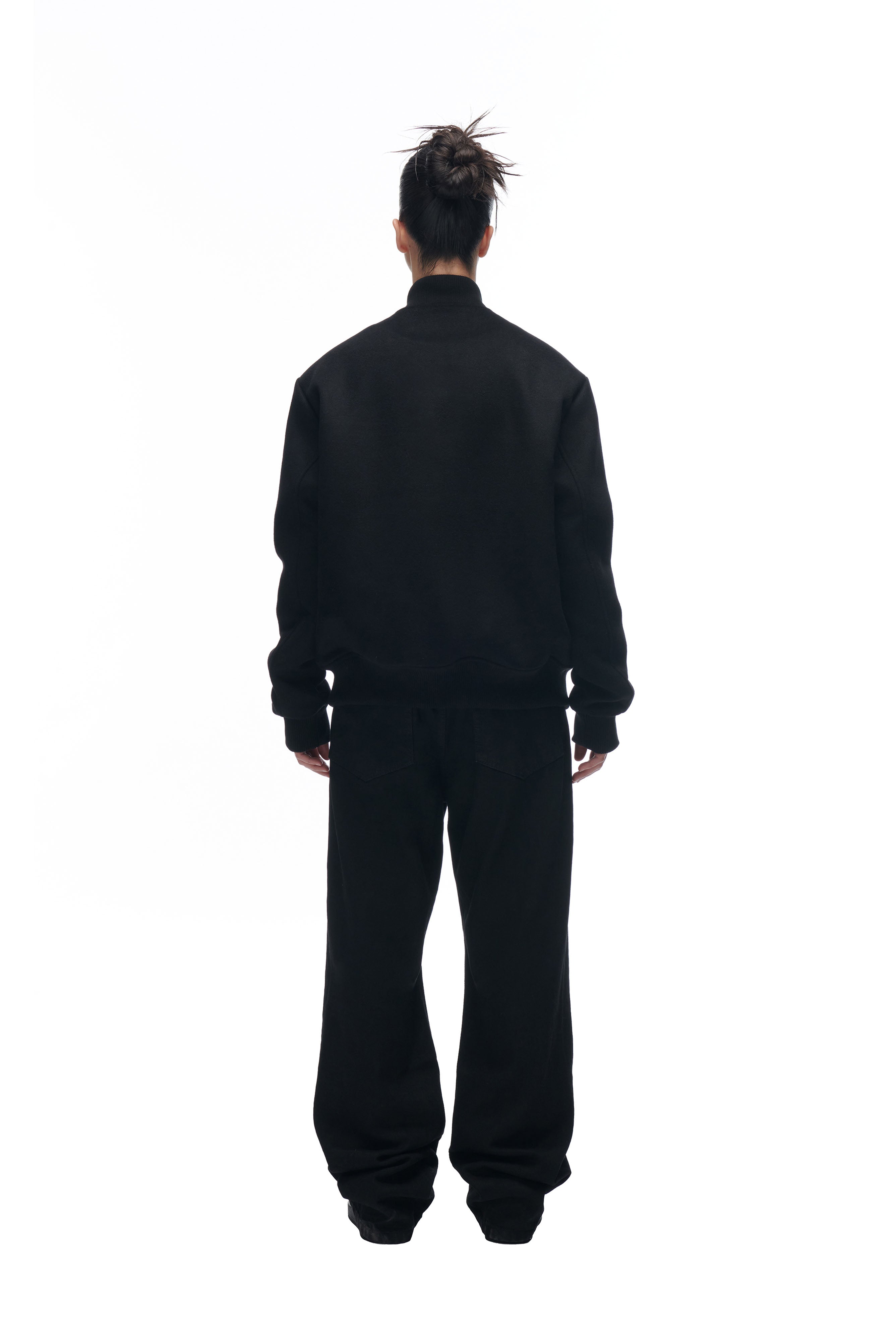 A person stands facing away from the camera, wearing the 6PM LH VARSITY JACKET and loose black pants, hair in a messy bun, against a plain white background.