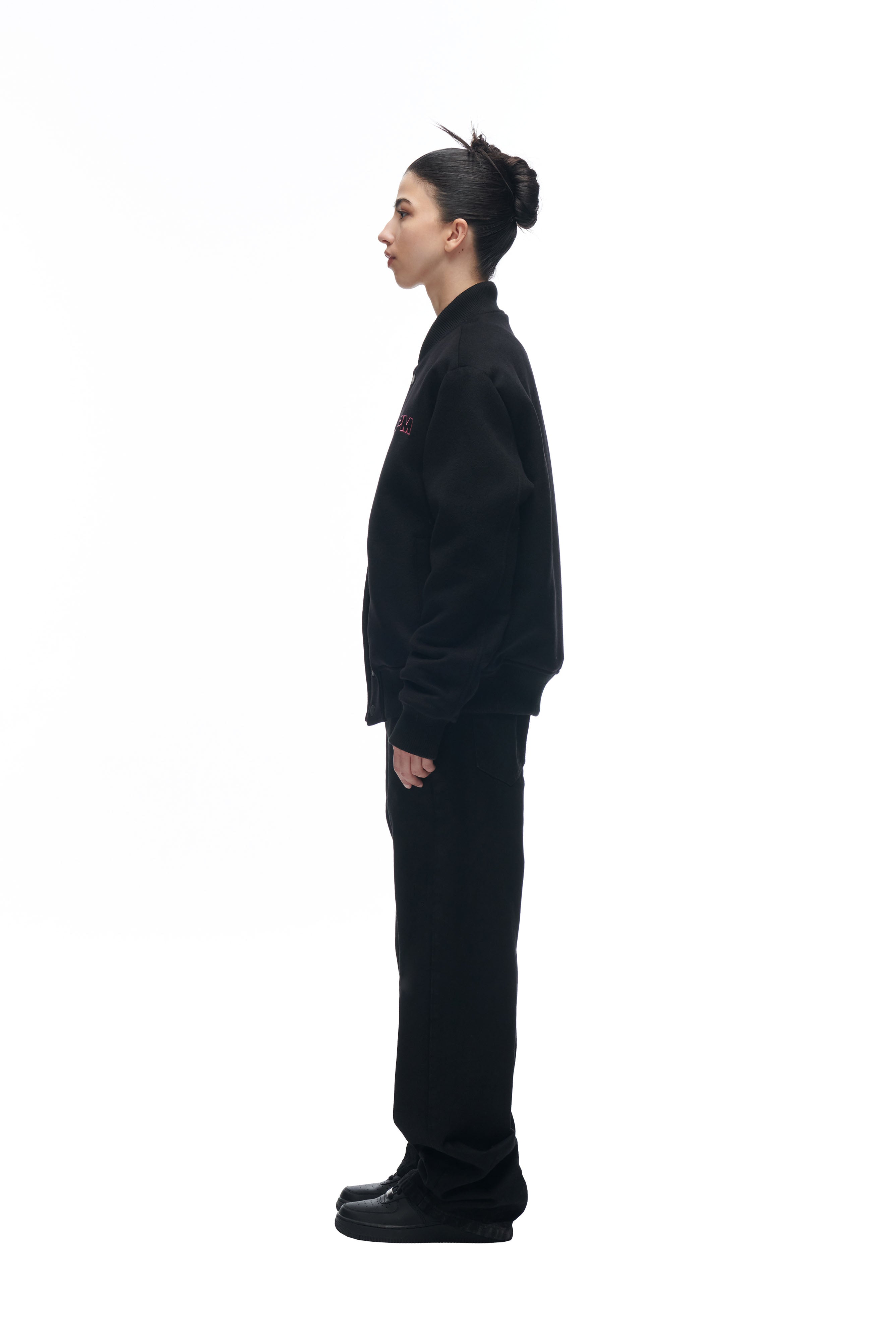 A person with dark hair in a bun stands in profile facing left, wearing the 6PM LH VARSITY JACKET with black pants and black shoes, against a plain white background.