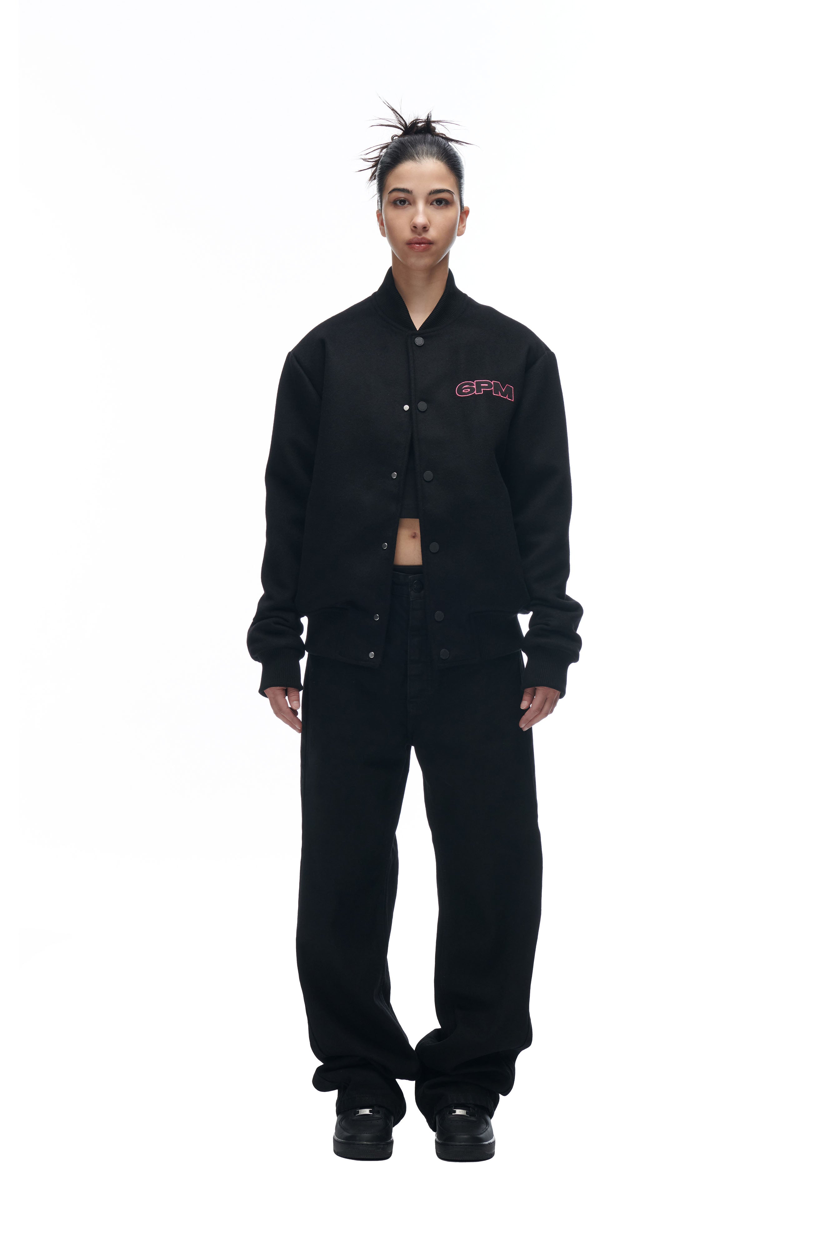 A person faces forward wearing the 6PM LH VARSITY JACKET with red “CFM” lettering, paired with loose black pants and black shoes. Their hair is in a messy bun, all set against a white background.