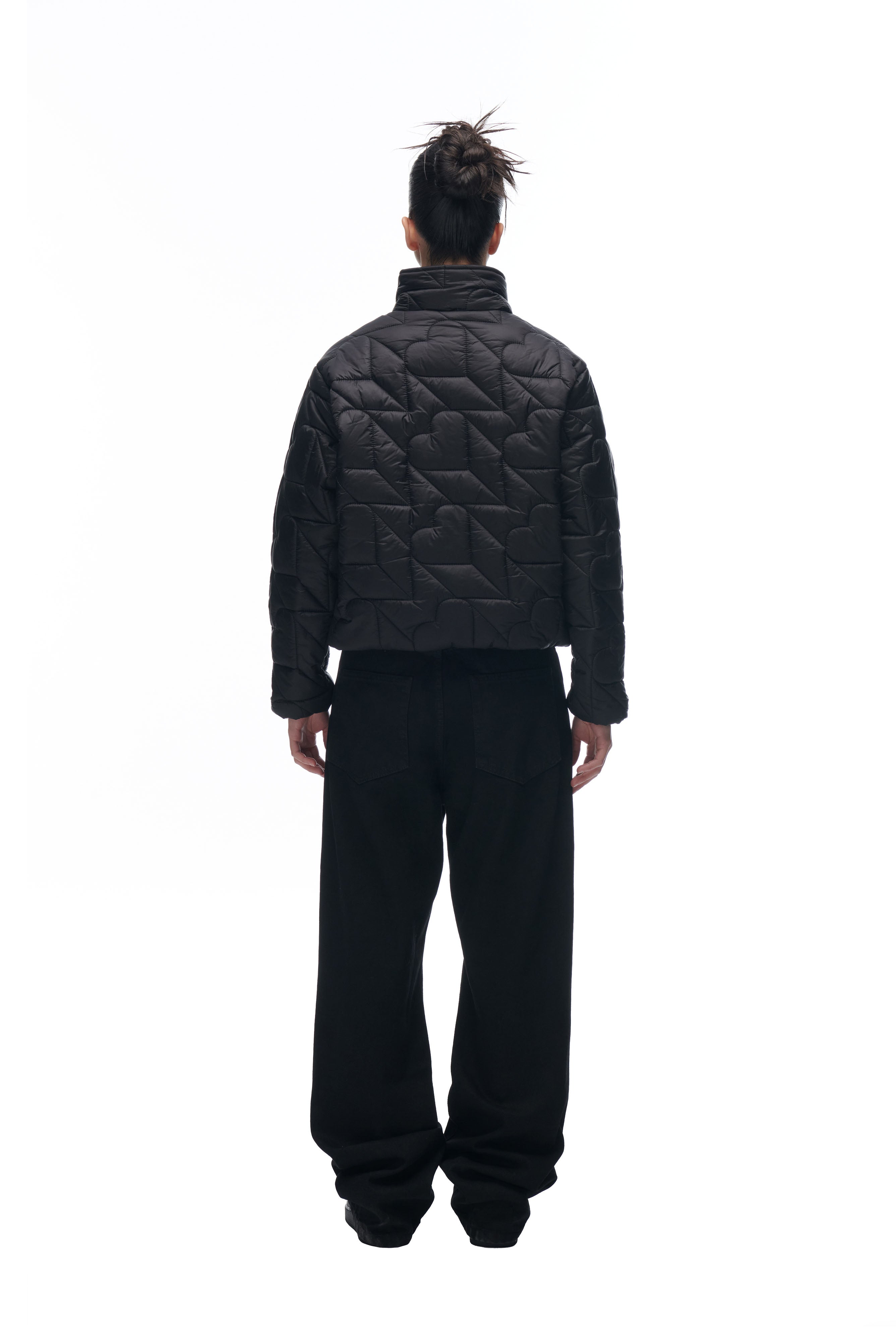 A person faces away, wearing the 6PM HEART SPRING JACKET—black and quilted—with loose black pants and hair in a messy bun, set against a plain white background.