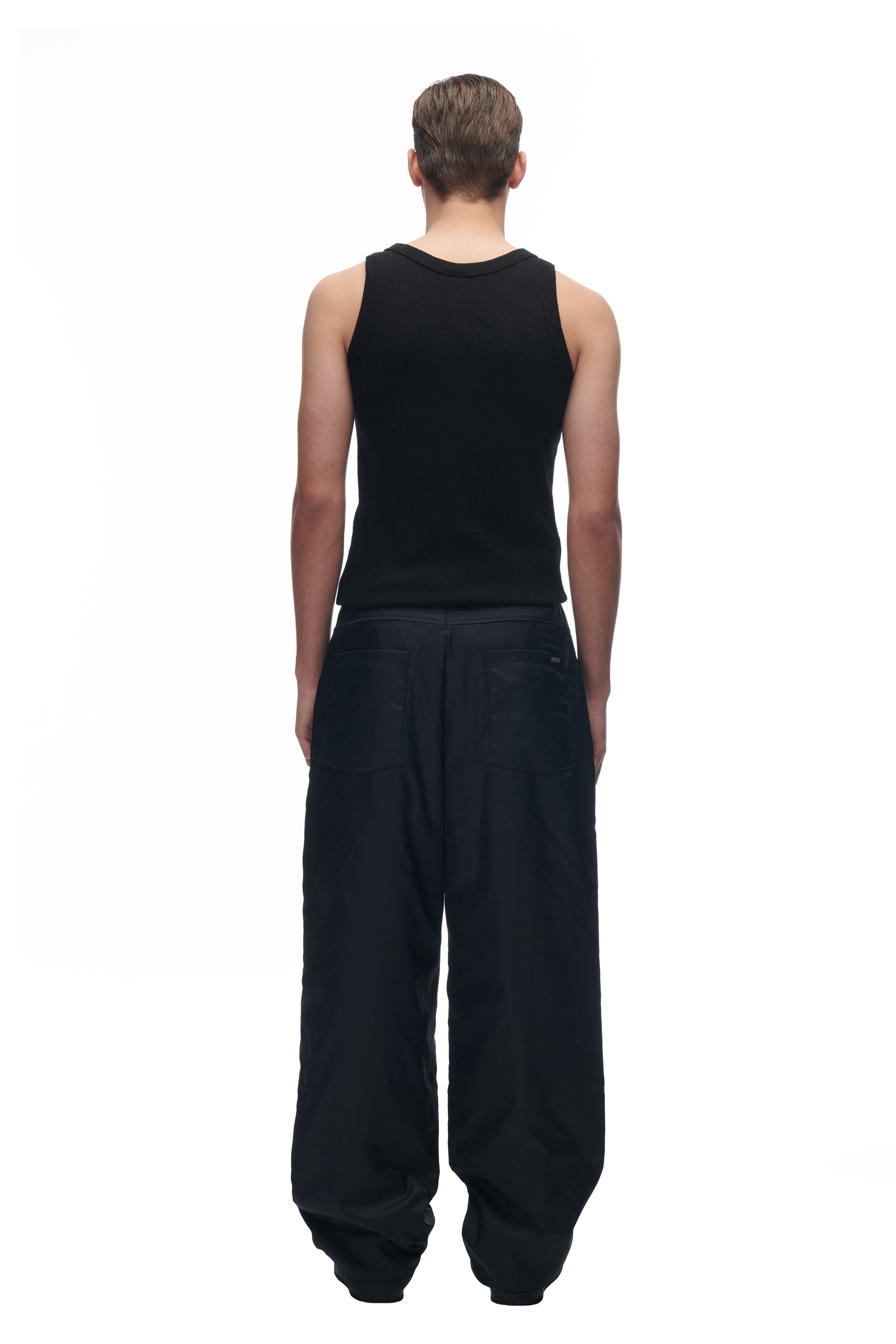 A person with short, slicked-back hair stands facing away from the camera against a plain white background, wearing 6PM’s black sleeveless top and LH NYLON PANTS BLACK.