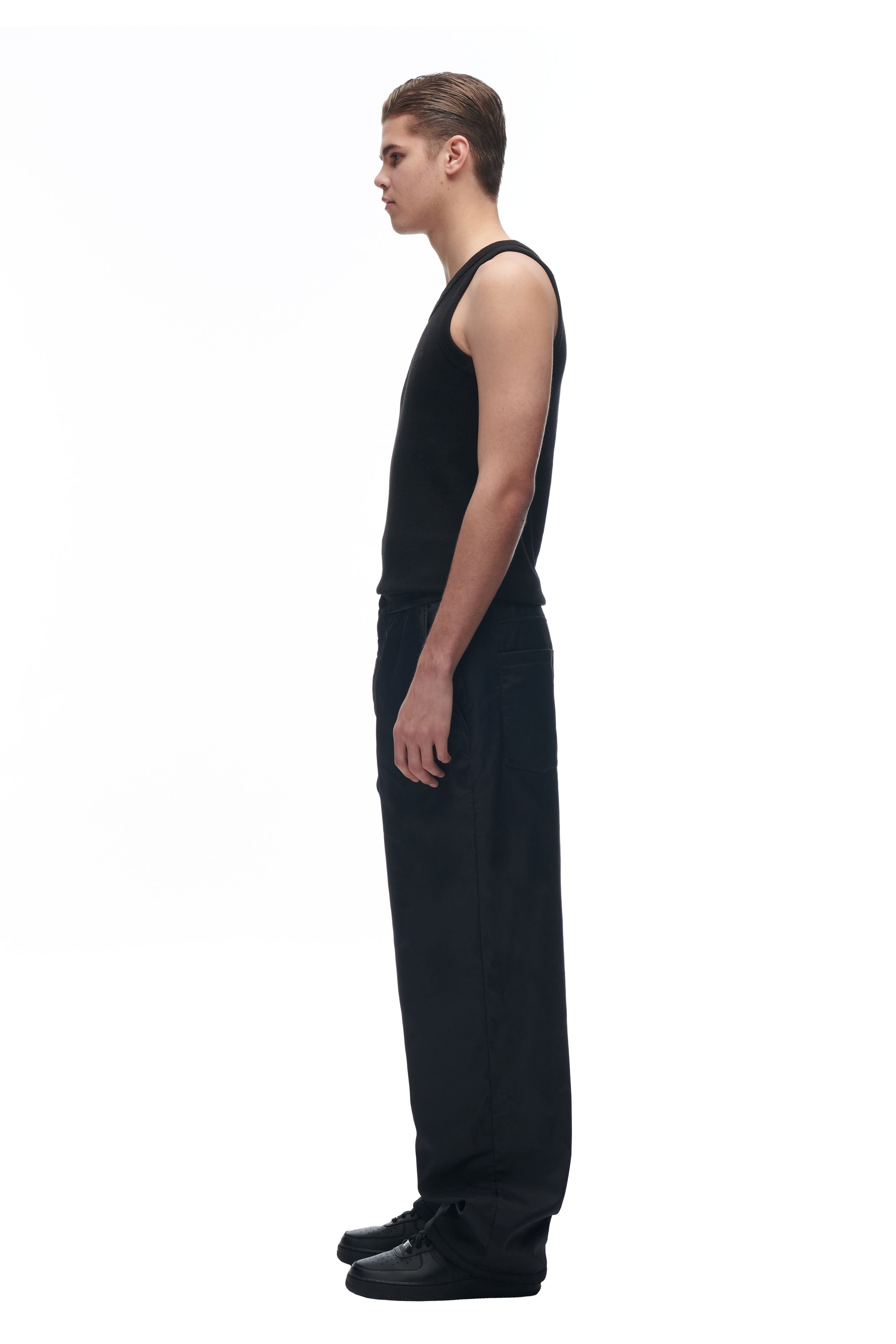 A person with short hair stands in profile facing left, wearing 6PM's LH NYLON PANTS BLACK along with a sleeveless black shirt and black shoes against a plain white background.
