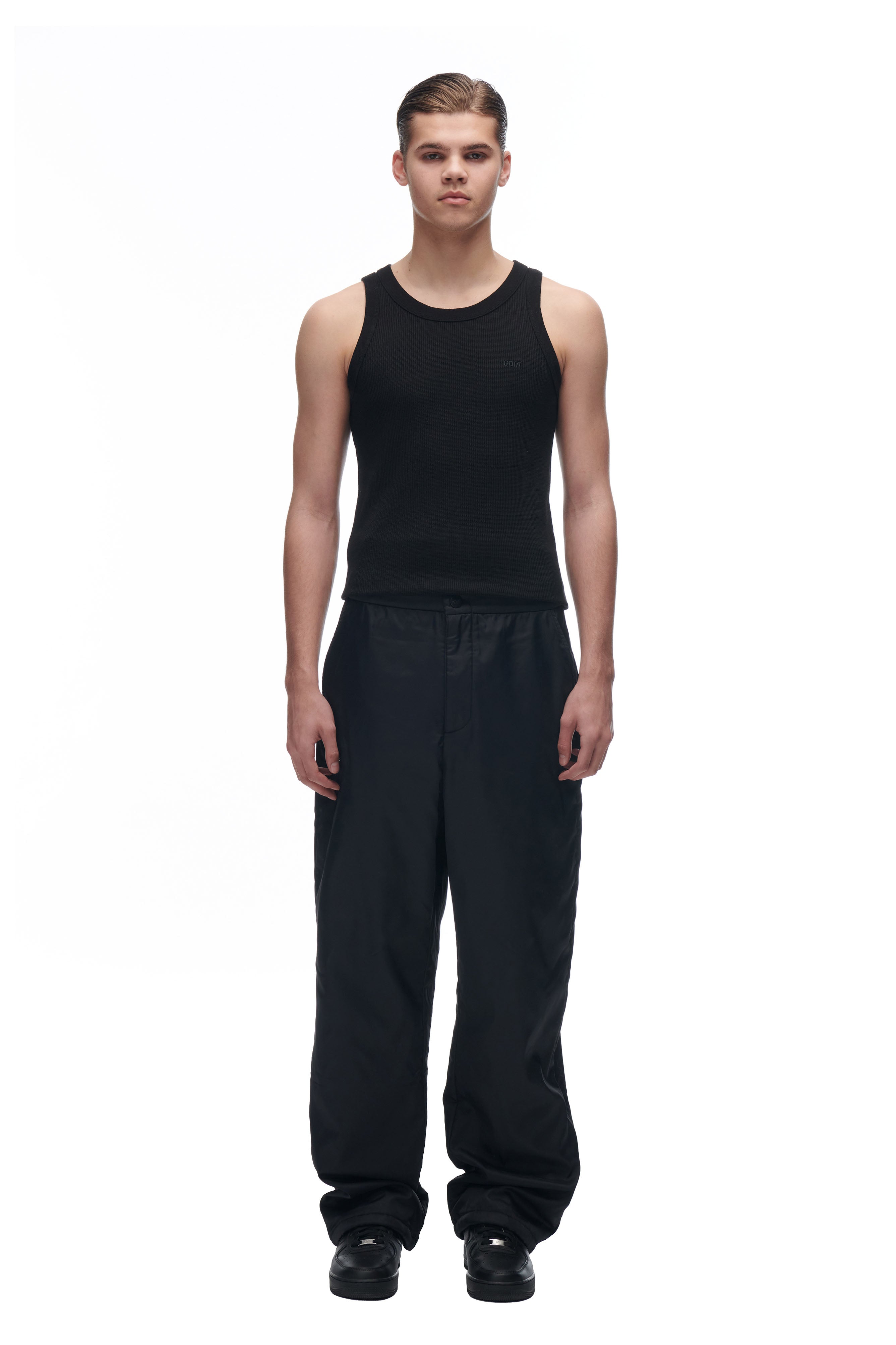 A young person stands facing forward against a plain white background, wearing 6PM’s LH NYLON PANTS BLACK with a fitted black tank top and black shoes, arms relaxed at their sides.
