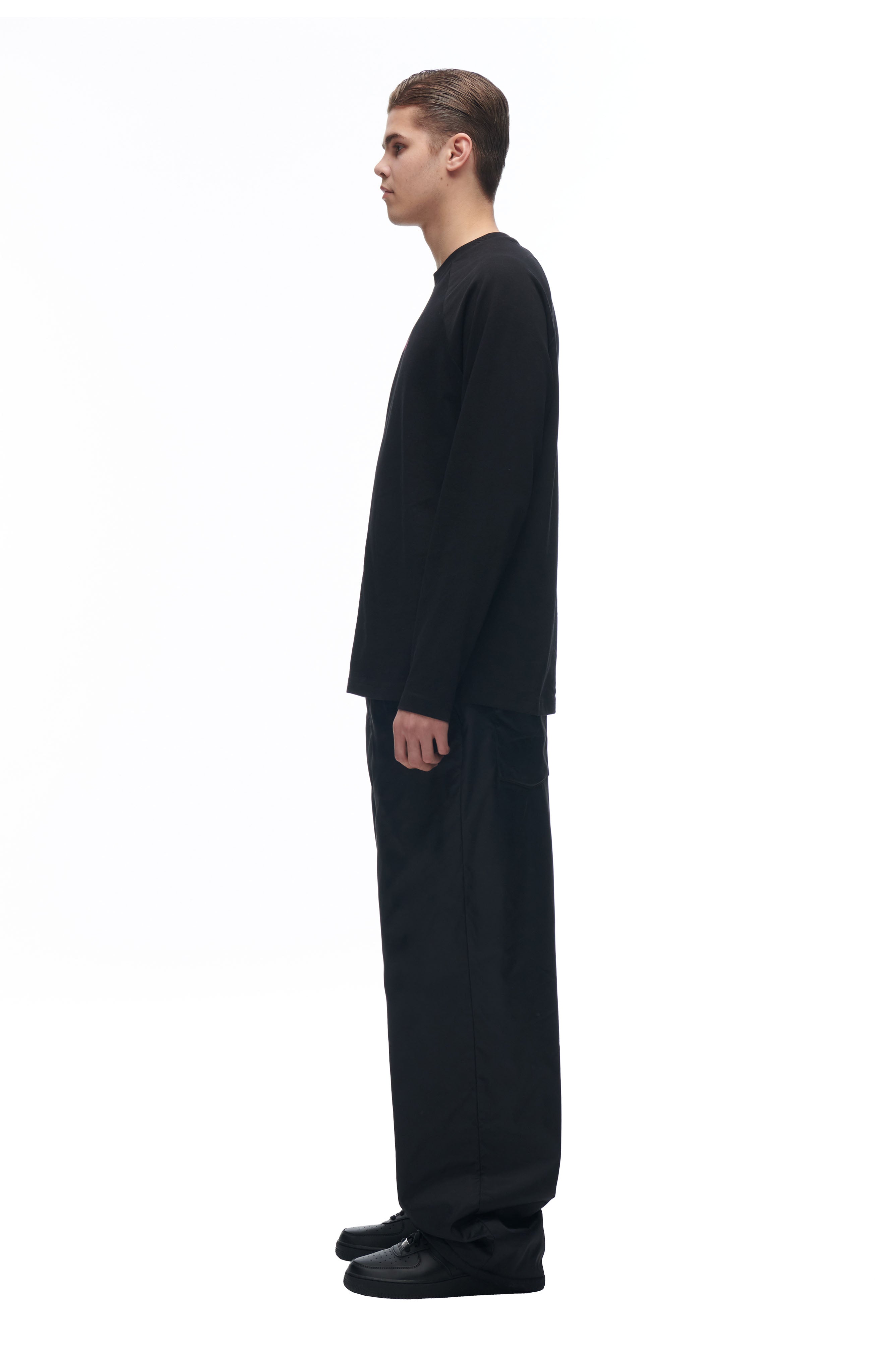 A person stands in profile against a white background wearing the 6PM LH LONGSLEEVE BLACK/BLACK shirt, paired with black pants and black shoes.
