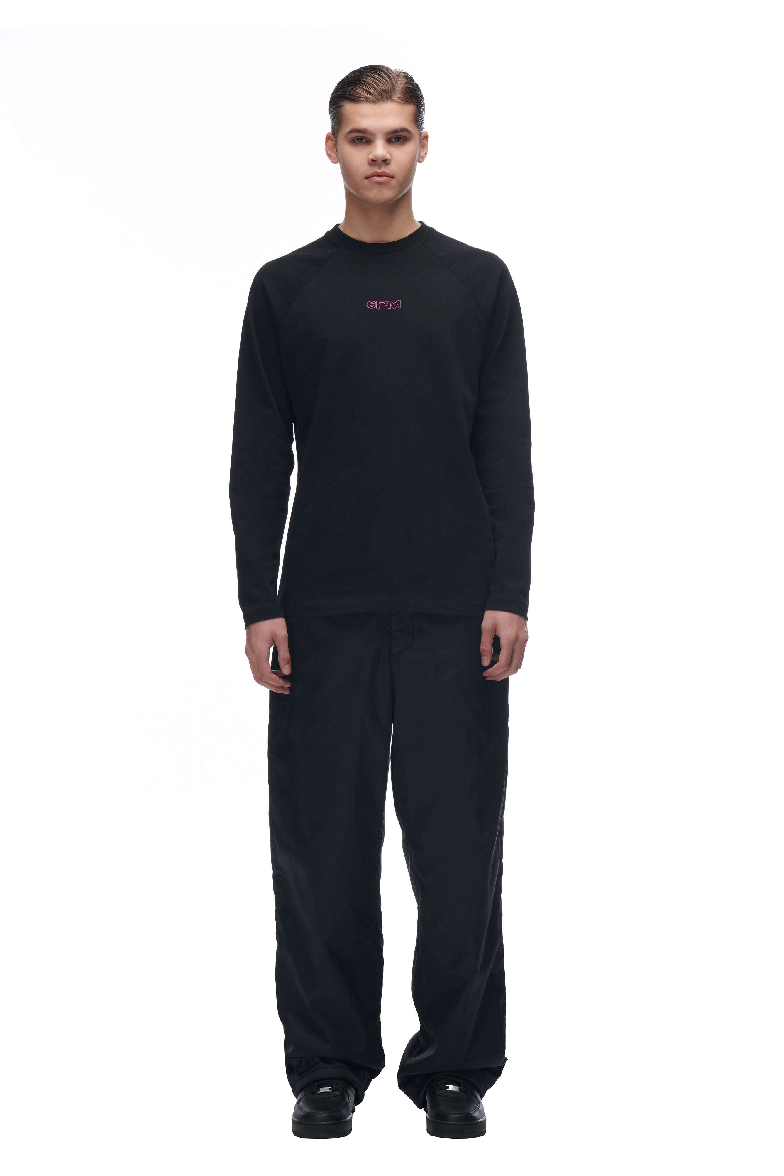 A person faces forward against a white background, wearing the 6PM LH LONGSLEEVE BLACK/BLACK shirt with loose black pants and black shoes. Their hair is neatly styled and they have a neutral expression.