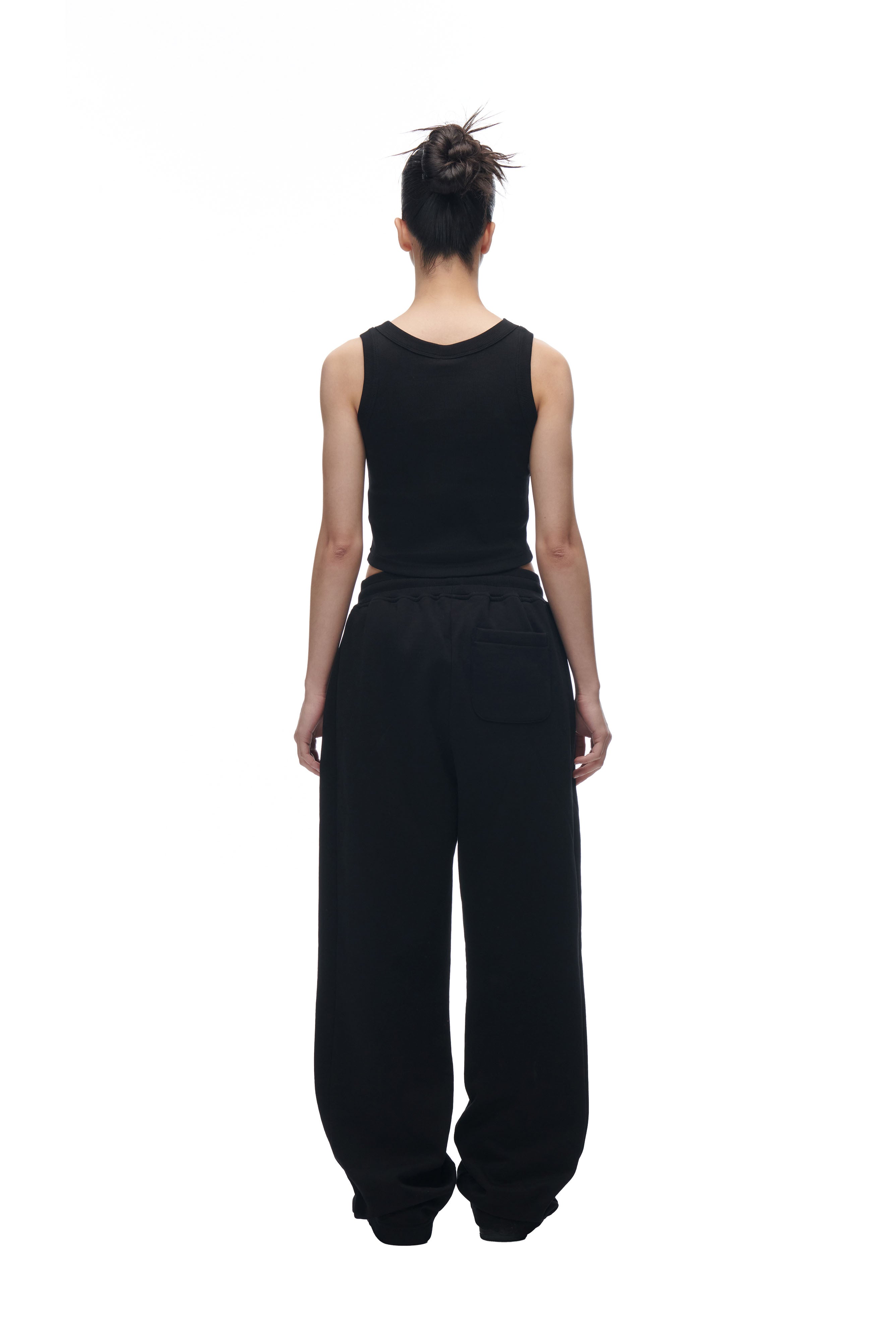 A person with their hair in a messy bun stands facing away from the camera, wearing a black sleeveless top and 6PM's LH SWEATPANT BLACK against a plain white background.