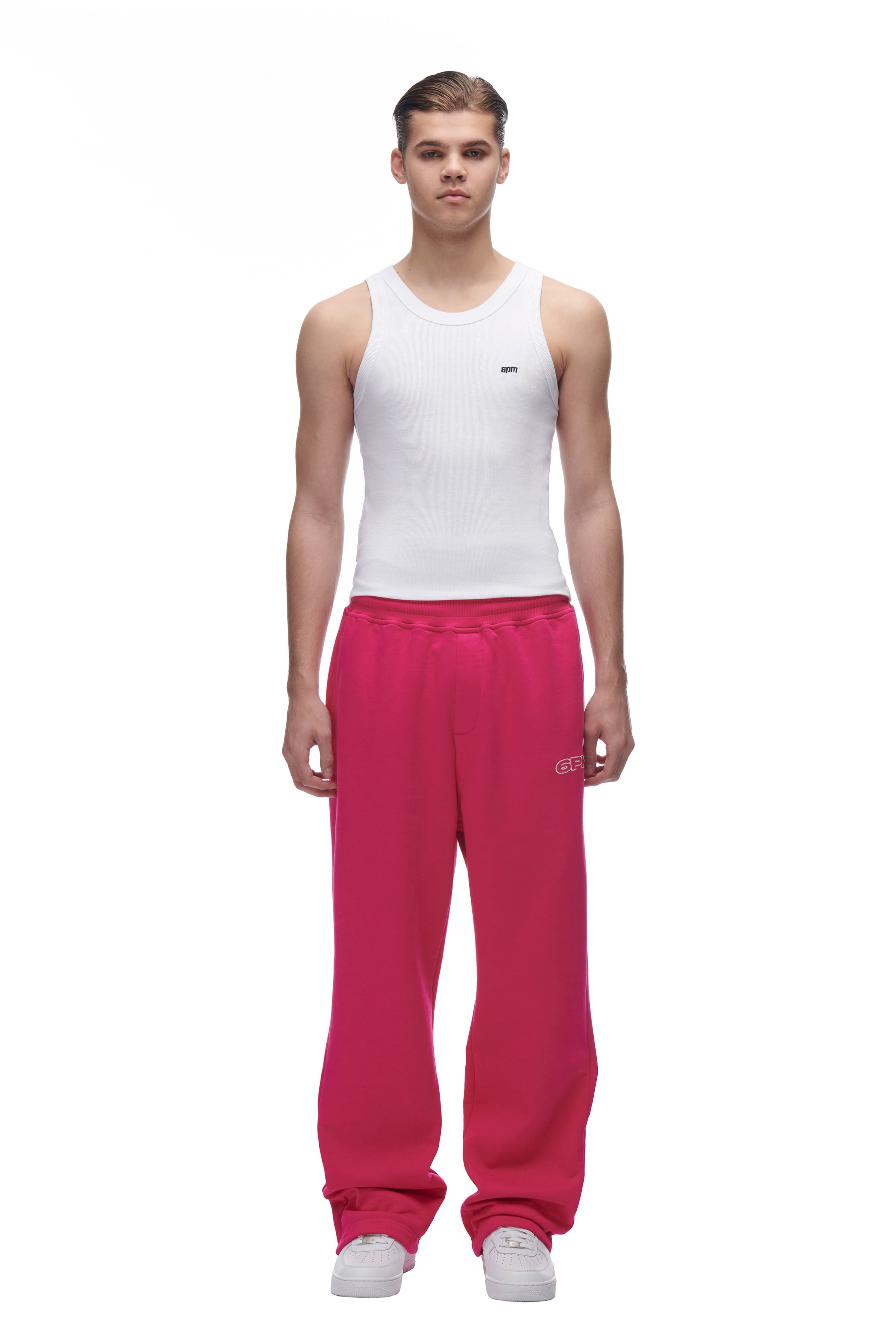 A person stands facing forward against a white background, wearing the 6PM LH SWEATPANT PINK, a white sleeveless tank top, and white sneakers.