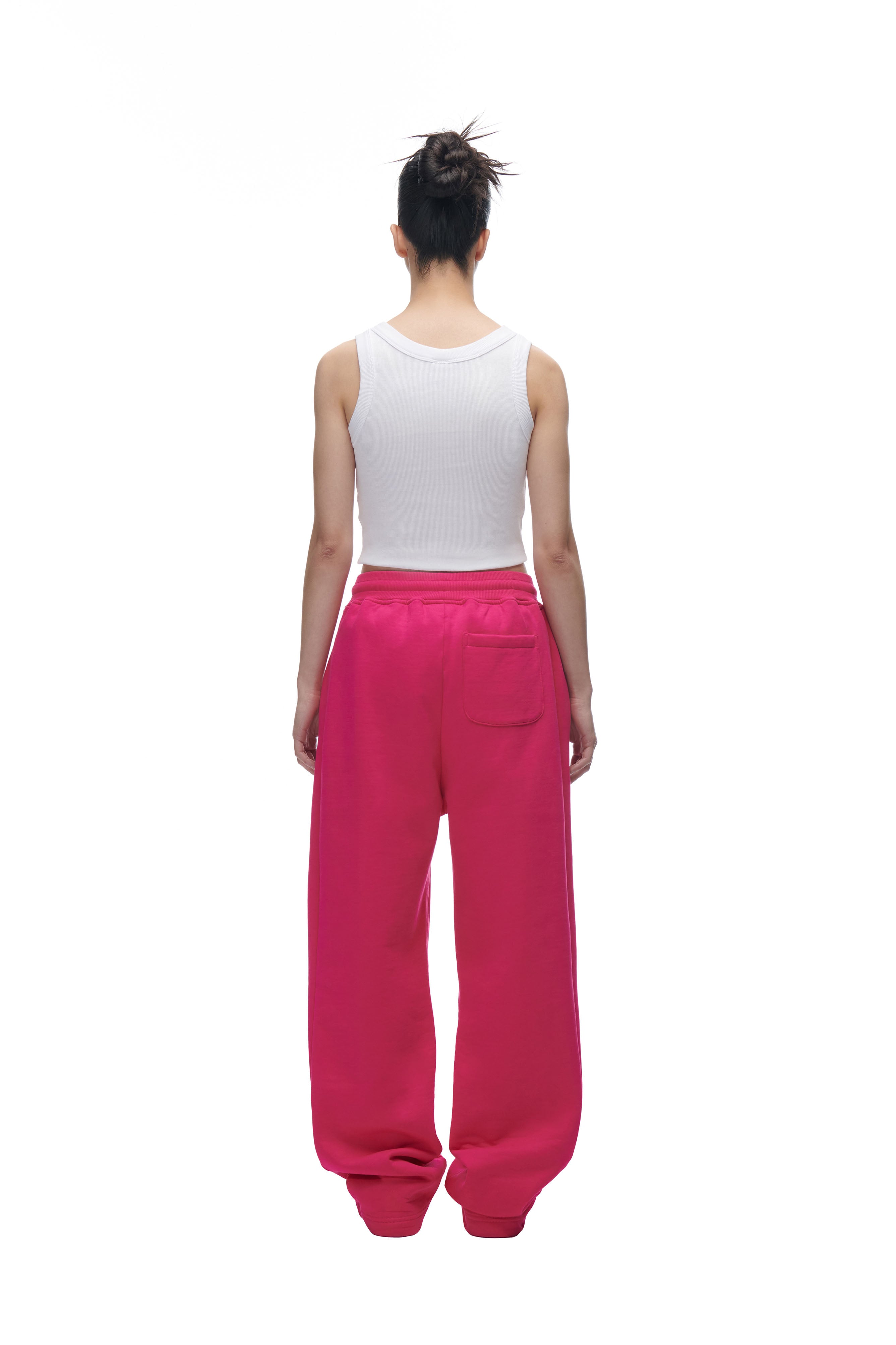 A person with a messy bun stands facing away against a plain white background, wearing a white sleeveless top and 6PM’s LH SWEATPANT PINK—loose, bright pink sweatpants with a back pocket.