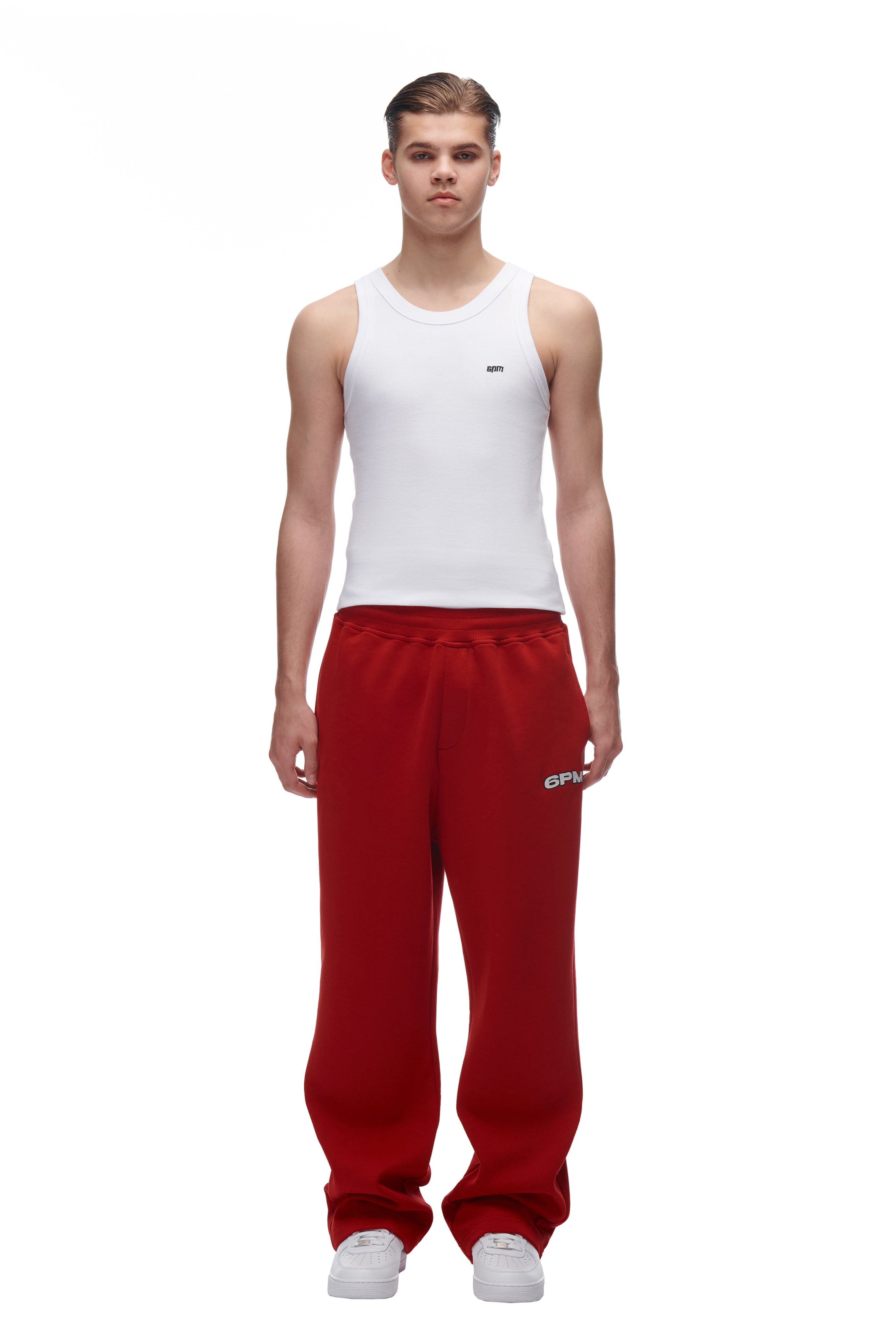 A young man faces forward against a plain white background, wearing 6PM's LH SWEATPANT RED, a fitted white tank top, and white sneakers.