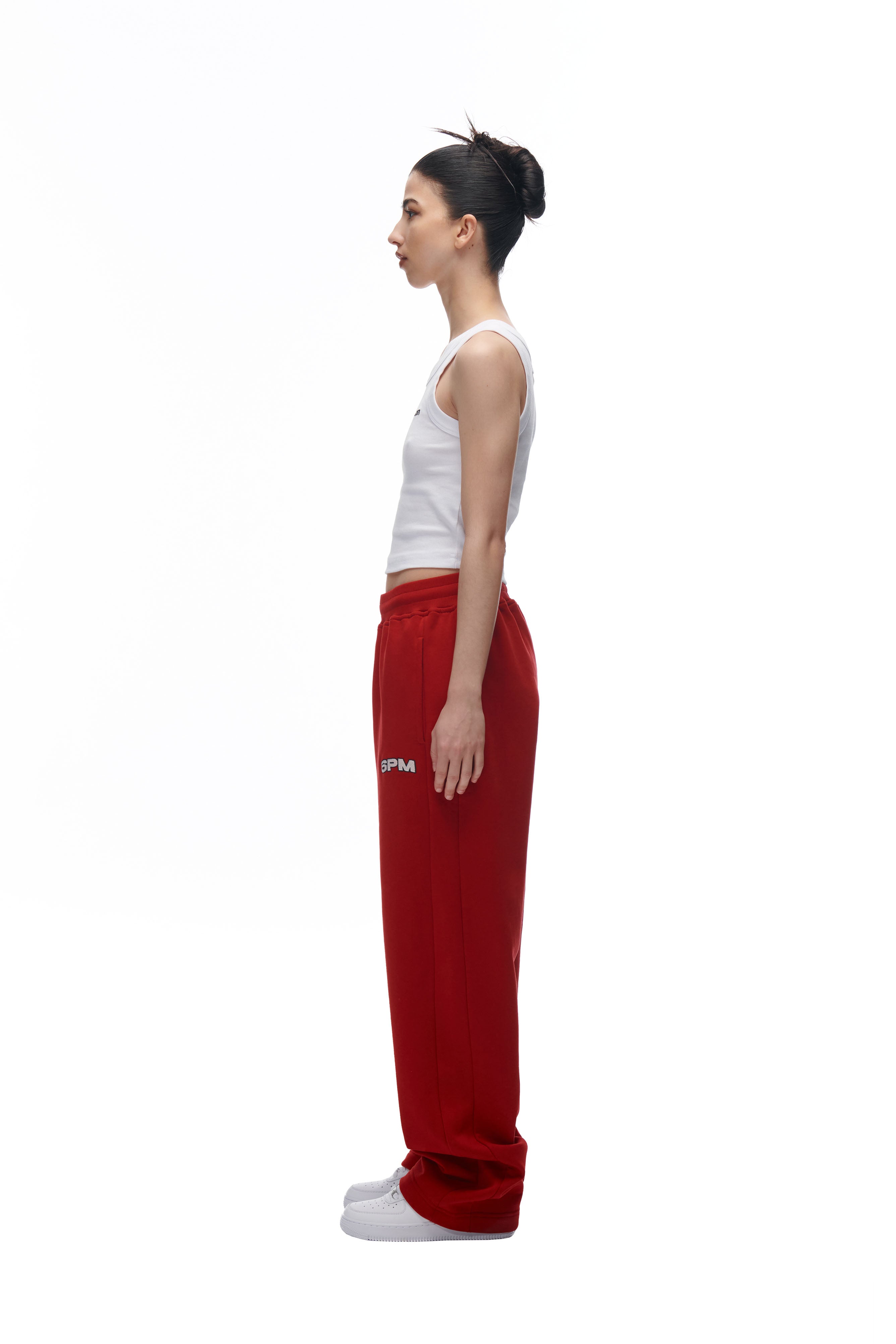 A woman stands in profile against a white background, wearing the 6PM LH SWEATPANT RED, a white tank top, and white sneakers. Her dark hair is tied up in a bun.