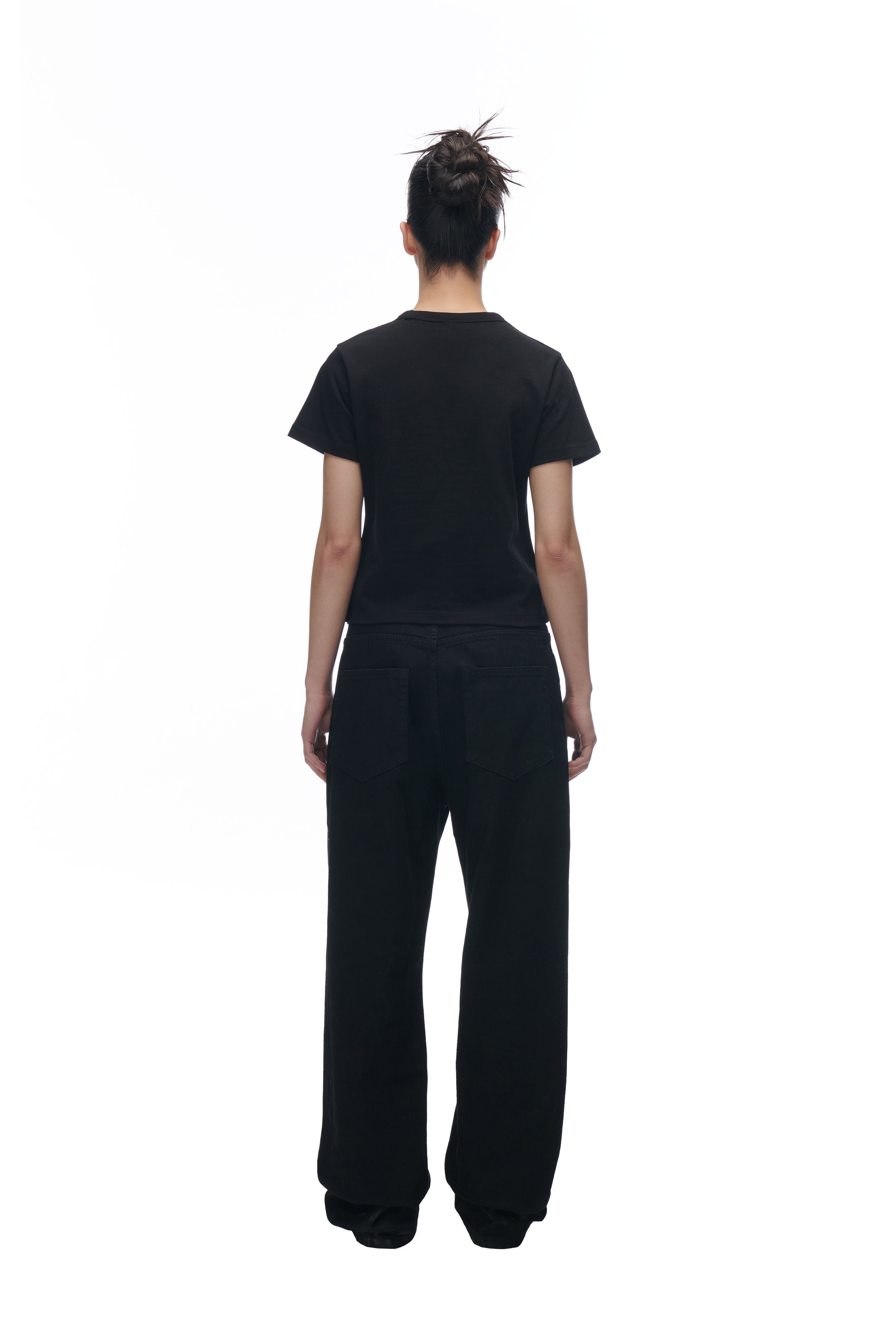 A person with dark hair in a messy bun wears the 6PM GIRLS BABYTEE BLACK, paired with loose black pants, and stands facing away against a white background.