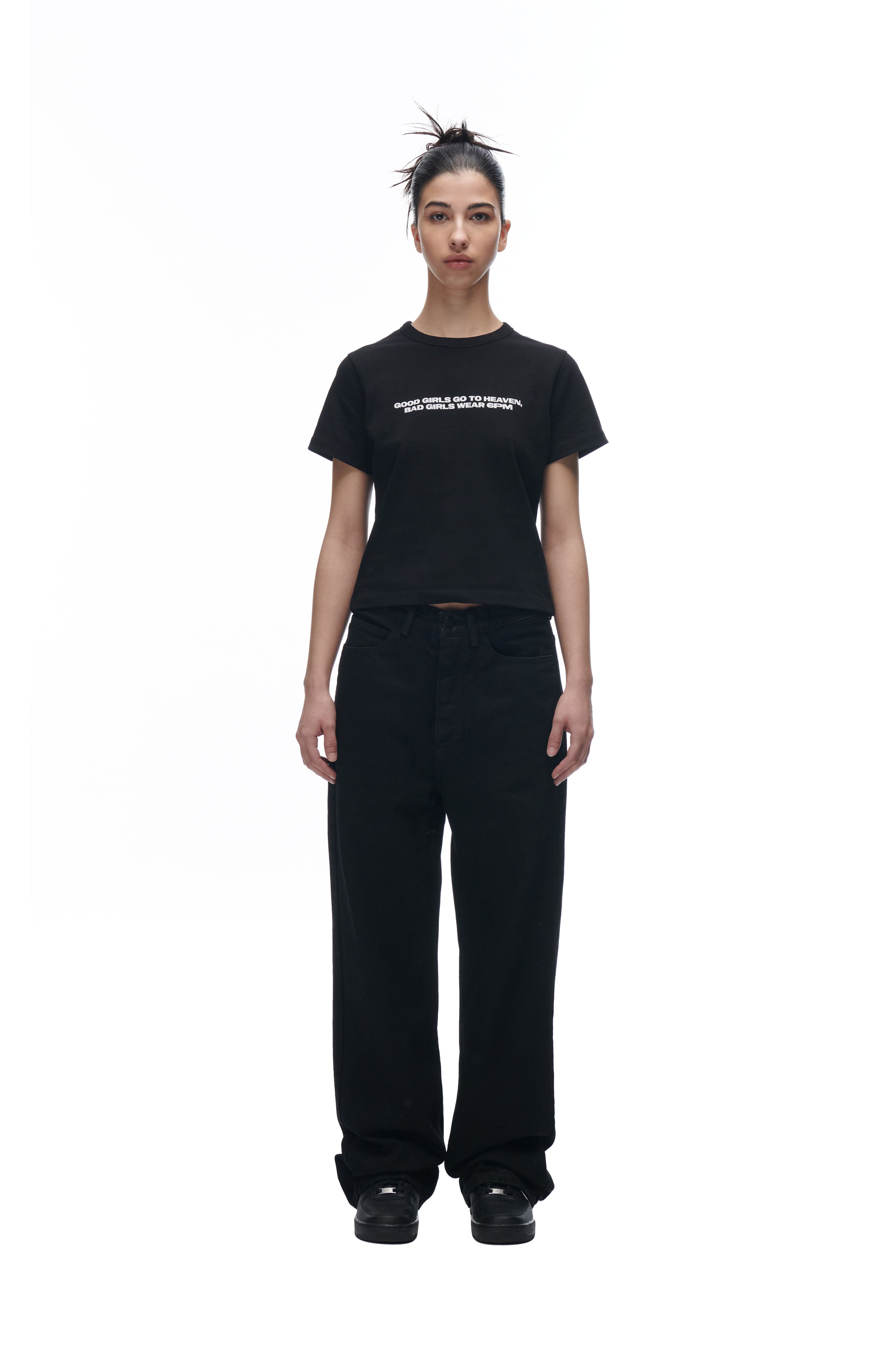 A person with dark hair in a messy bun stands facing forward against a plain white background, wearing the 6PM GIRLS BABYTEE BLACK with white text, paired with loose black pants and black shoes.