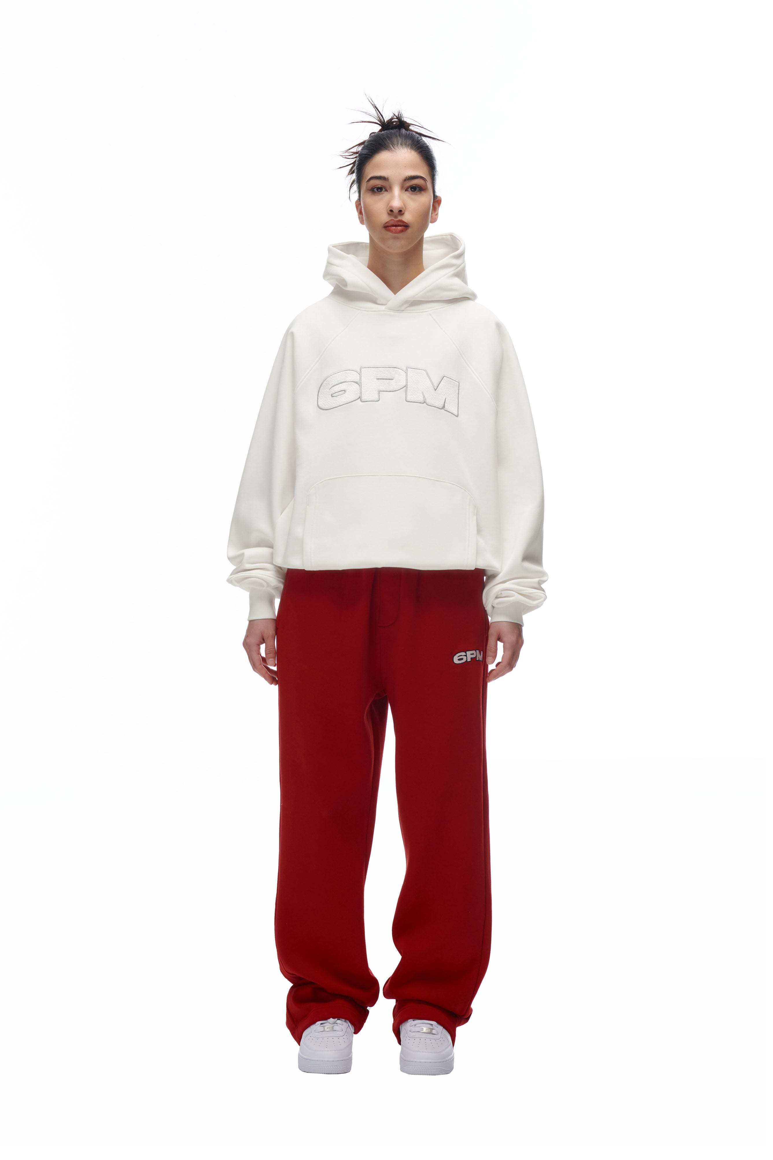 A person stands facing forward, wearing the LH HOODIE OFFWHITE by 6PM, red sweatpants with a small 6PM logo, and white sneakers. Their hair is in a messy bun against a plain white background.