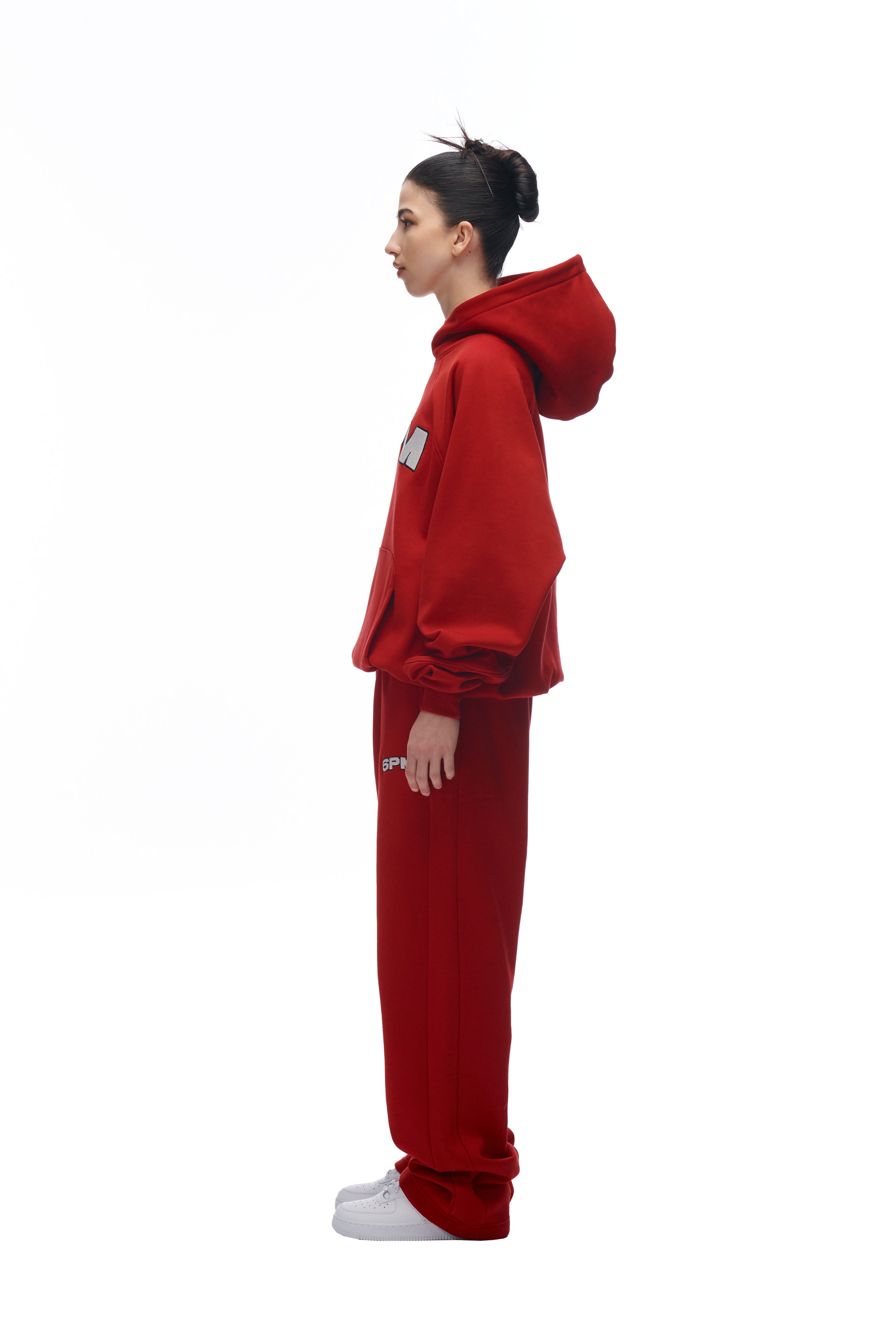 A woman stands in profile against a white background, wearing the 6PM LH HOODIE RED with a hood, matching red sweatpants, and white sneakers. Her hair is pulled up into a bun.
