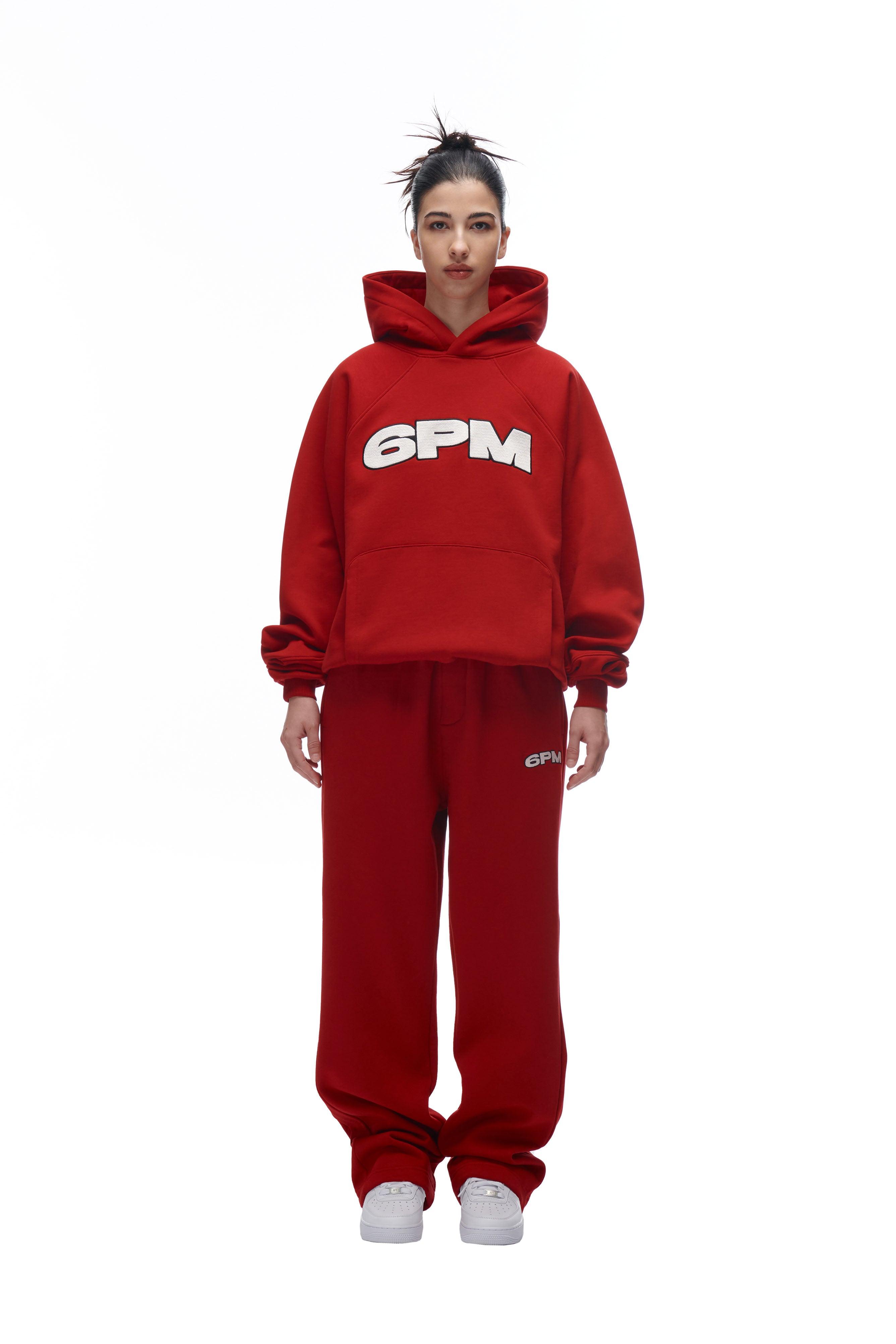 A person stands facing forward, wearing the LH HOODIE RED and matching sweatpants by 6PM, both featuring white 6PM branding. They have white sneakers, hair in a messy bun, and the background is plain white.