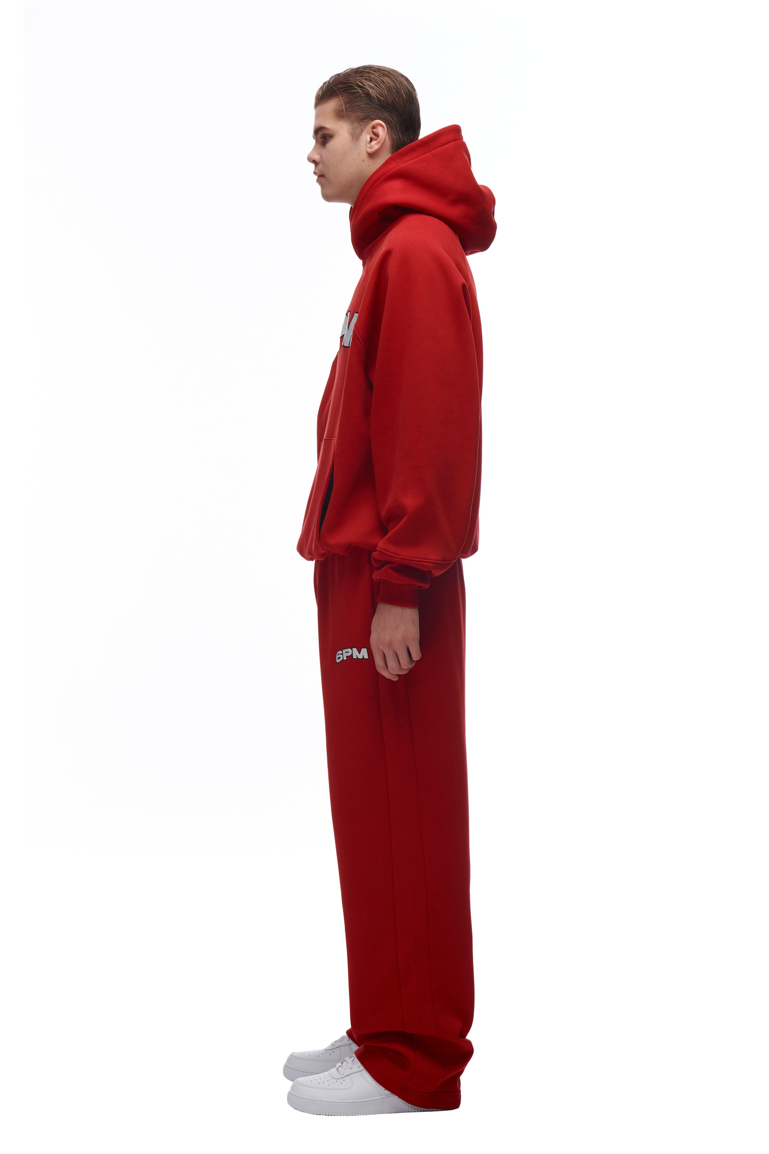 A person stands in profile against a white background, wearing a matching 6PM LH HOODIE RED and sweatpants with white text, paired with white sneakers.