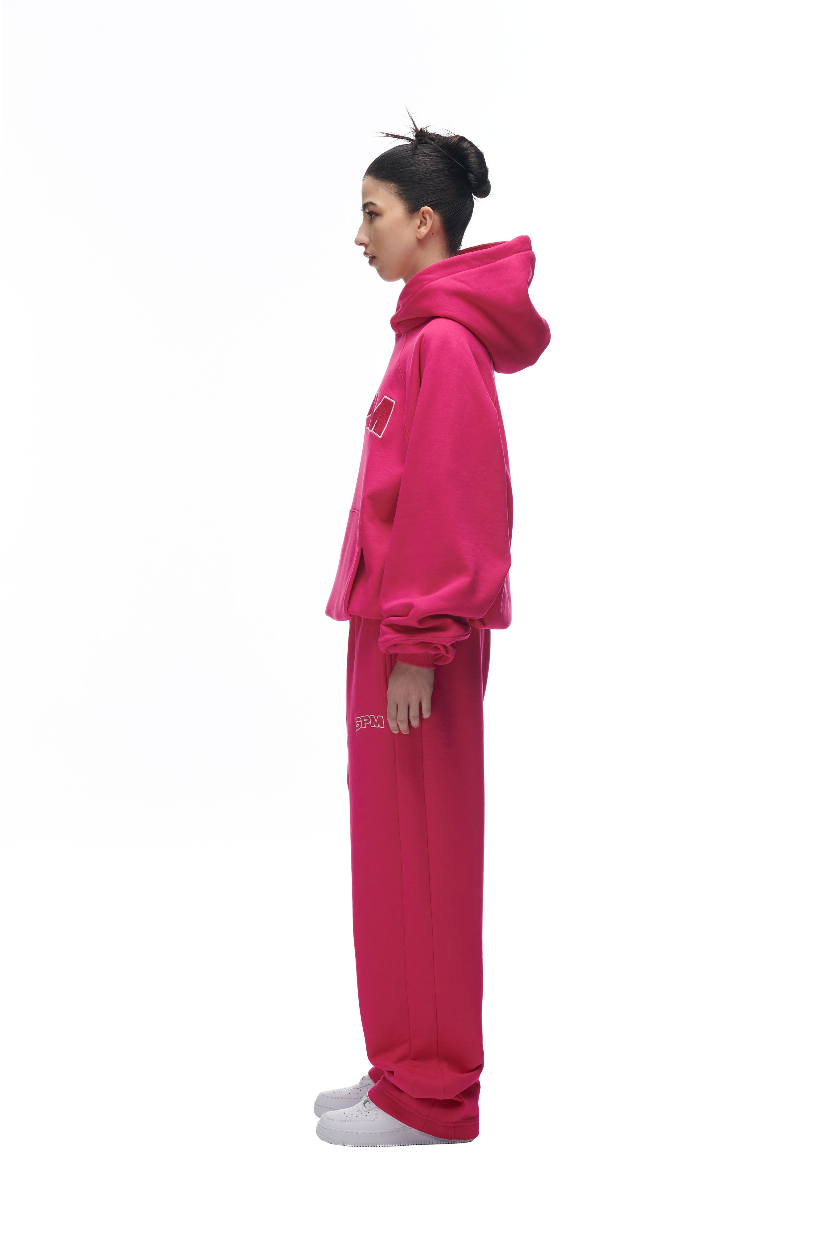 A woman in profile stands against a white background wearing the 6PM LH HOODIE PINK with matching sweatpants. Her dark hair is in a bun and she pairs the look with white socks.