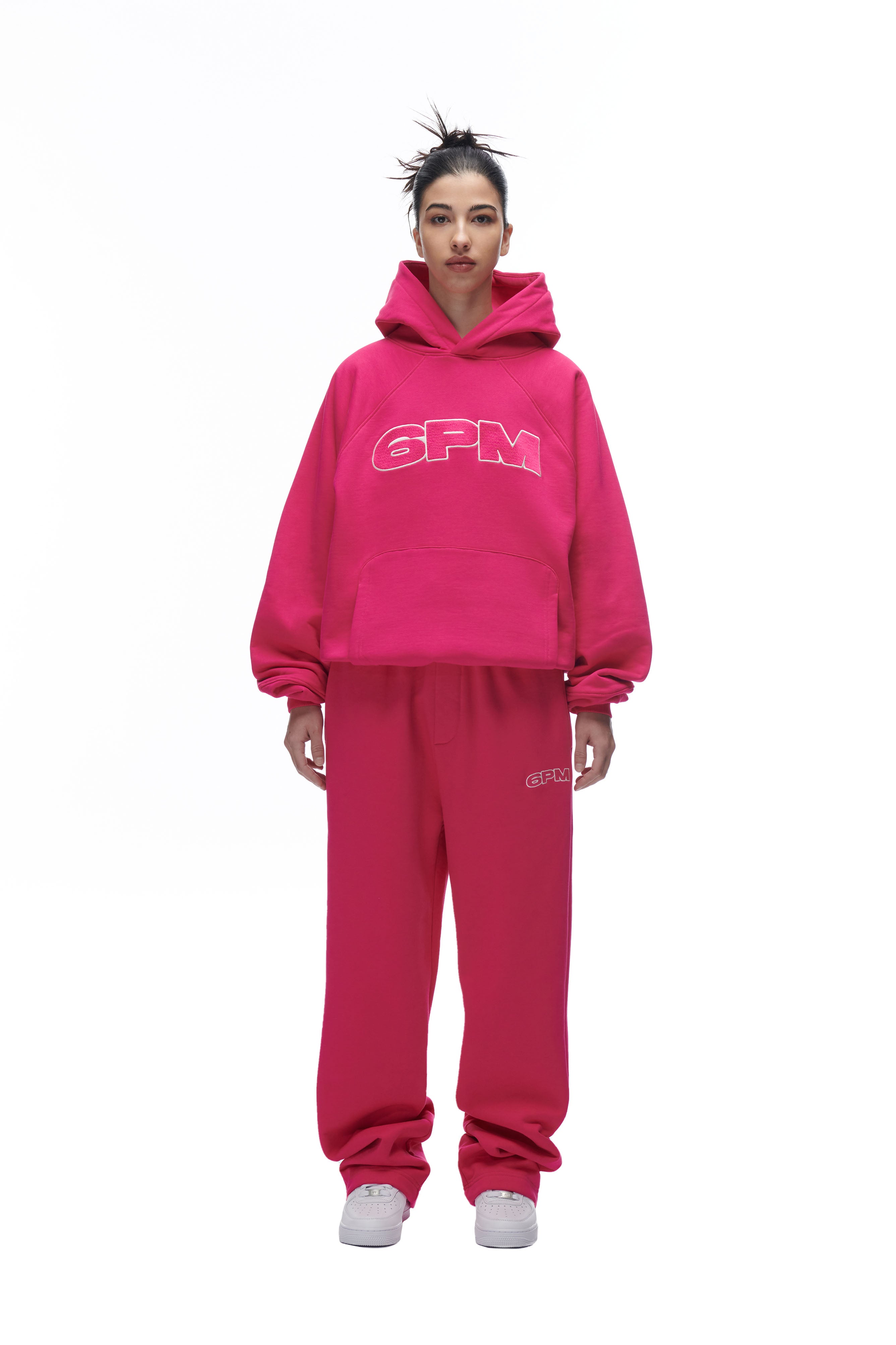 A person stands against a white background wearing the 6PM LH HOODIE PINK with matching pink sweatpants and white sneakers. Their hair is styled in a high, messy bun.
