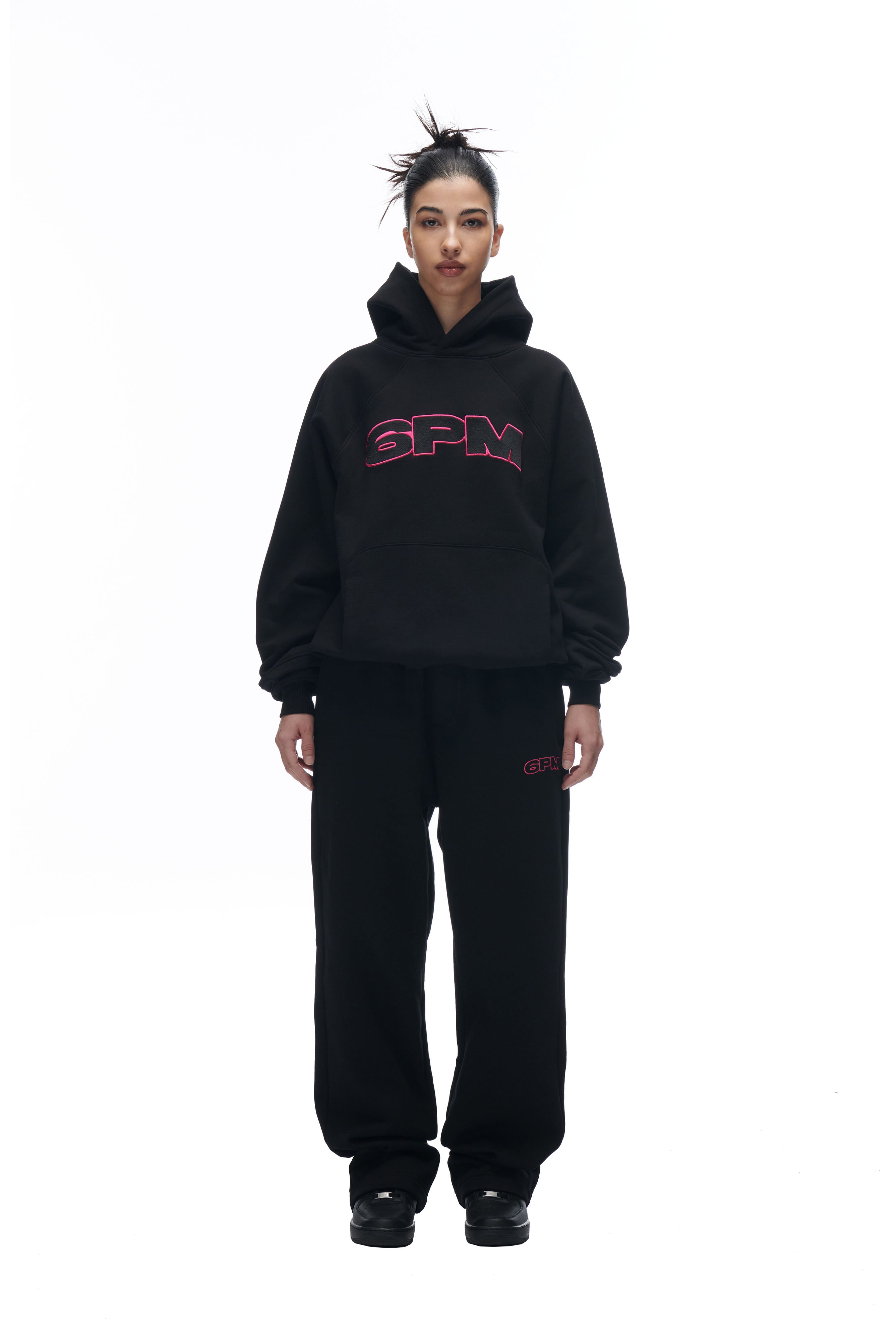 A person with a messy bun and neutral expression wears the 6PM LH HOODIE BLACK with matching black sweatpants, standing against a white background.