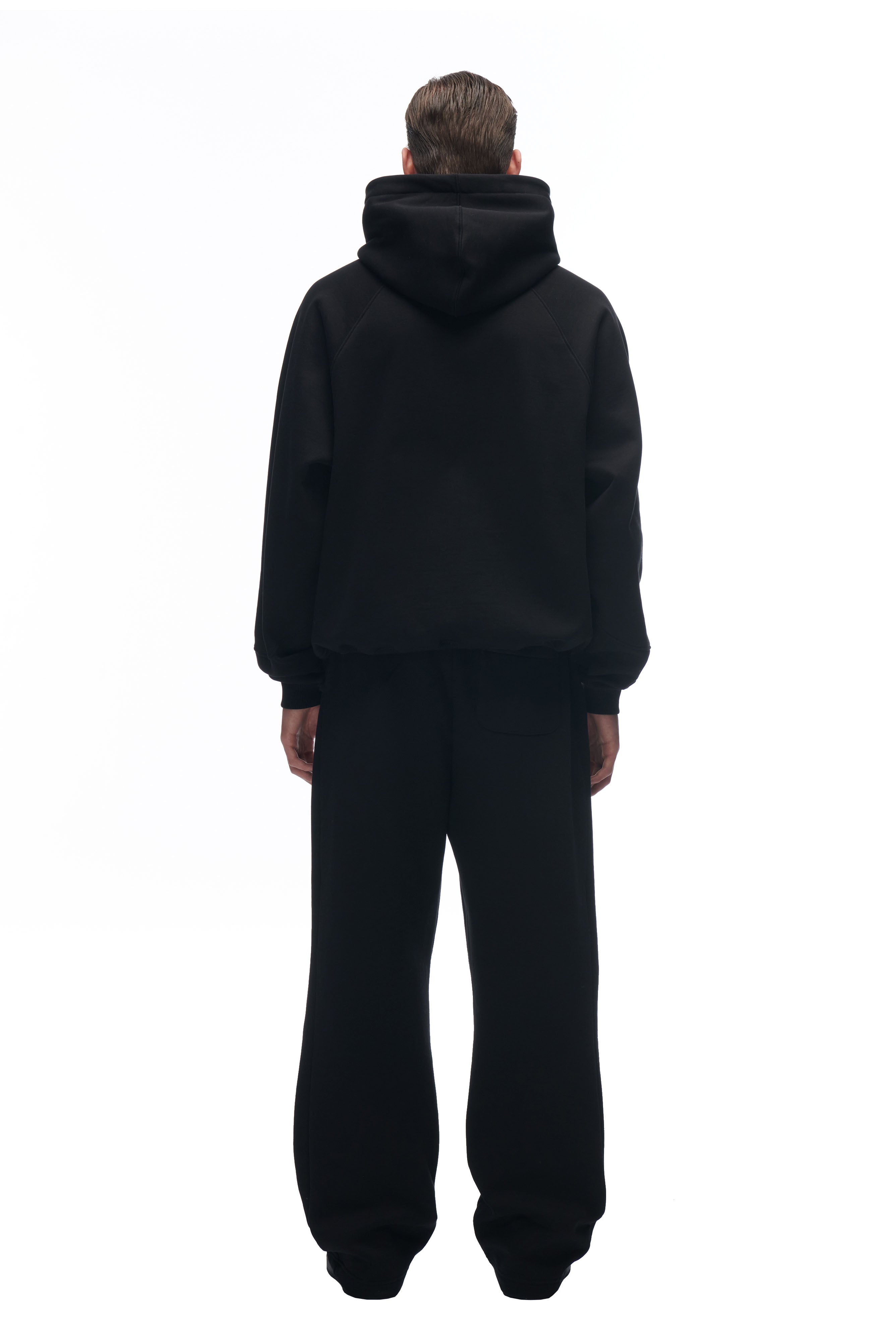 A person faces away, wearing the 6PM LH HOODIE BLACK with matching black pants. The plain white background emphasizes the minimalist, monochrome look.