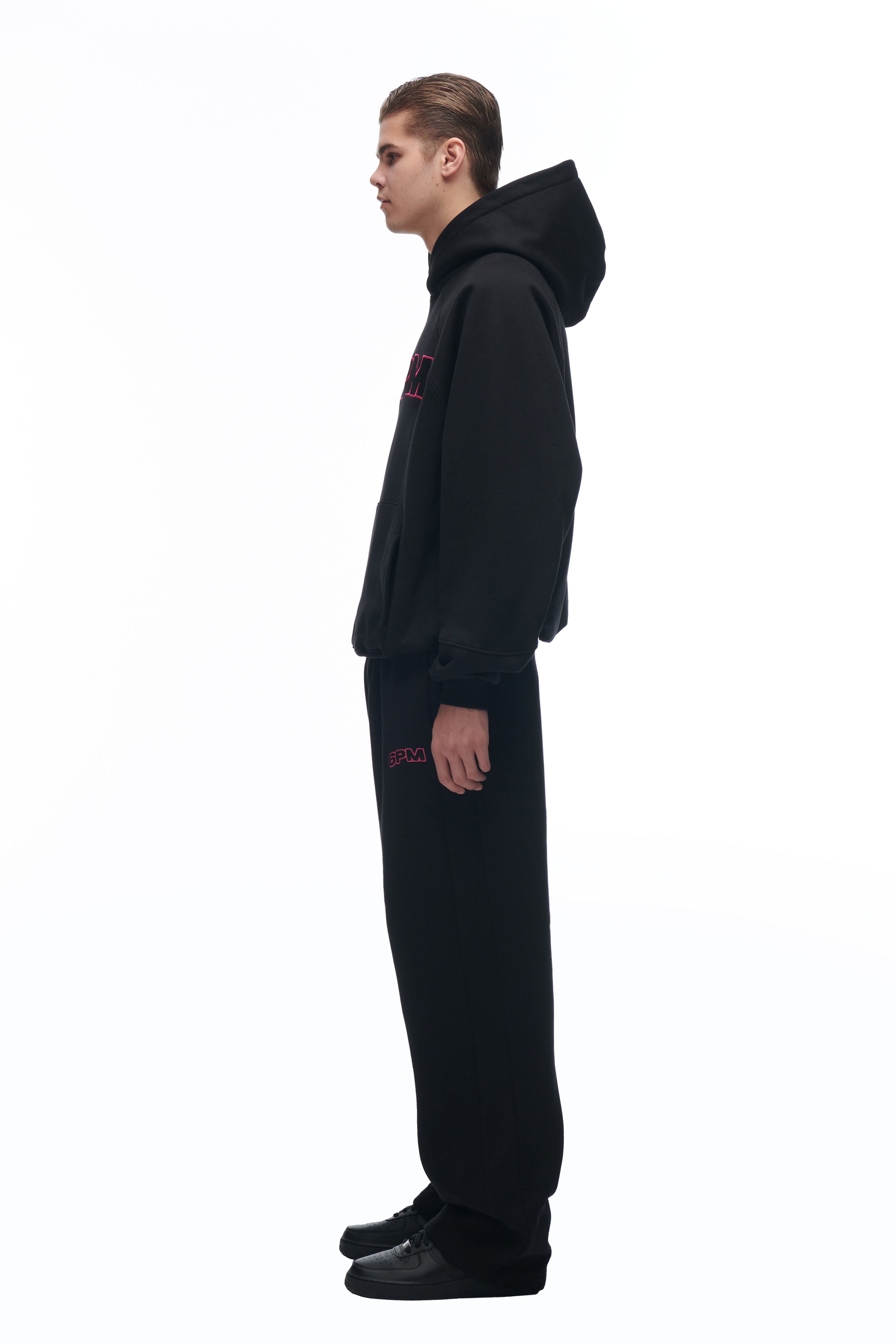 A person stands in profile against a white background, wearing the 6PM LH HOODIE BLACK with the hood down, paired with black sweatpants and black sneakers.