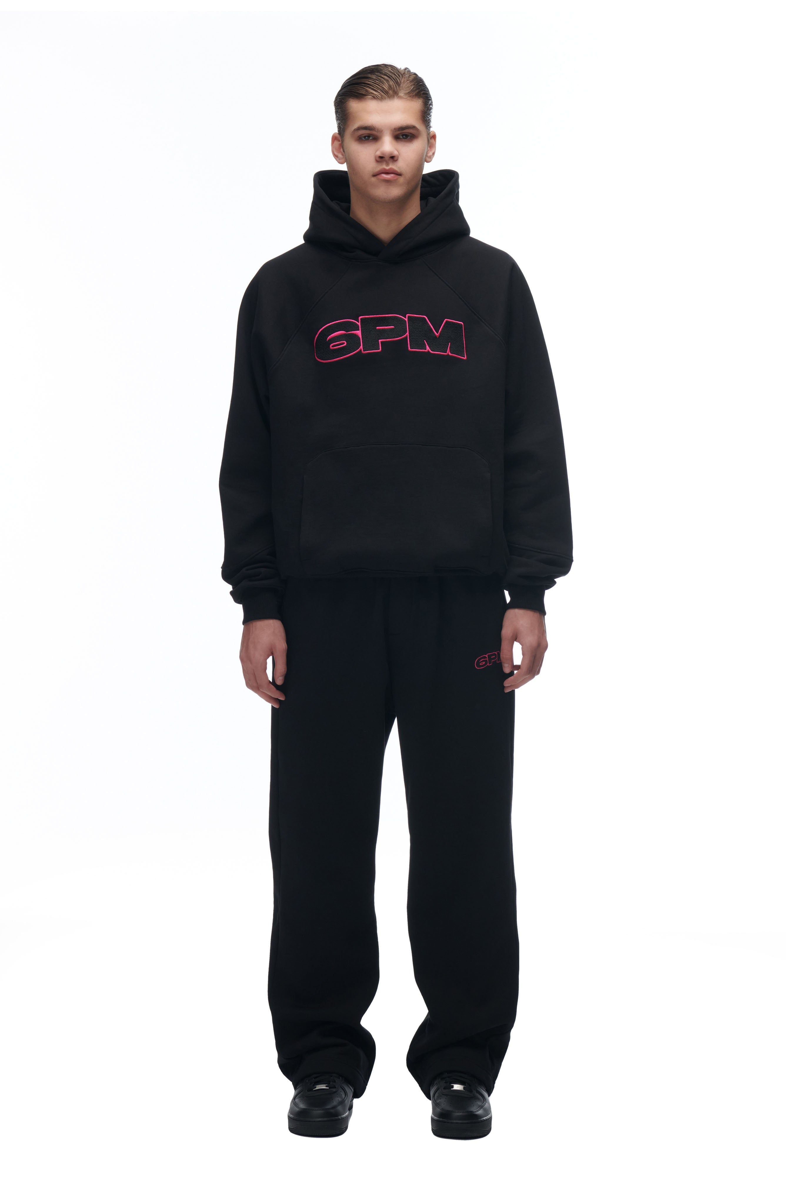 A person faces forward wearing the LH HOODIE BLACK by 6PM, featuring pink 6PM lettering, paired with matching black sweatpants and black shoes, set against a white background.